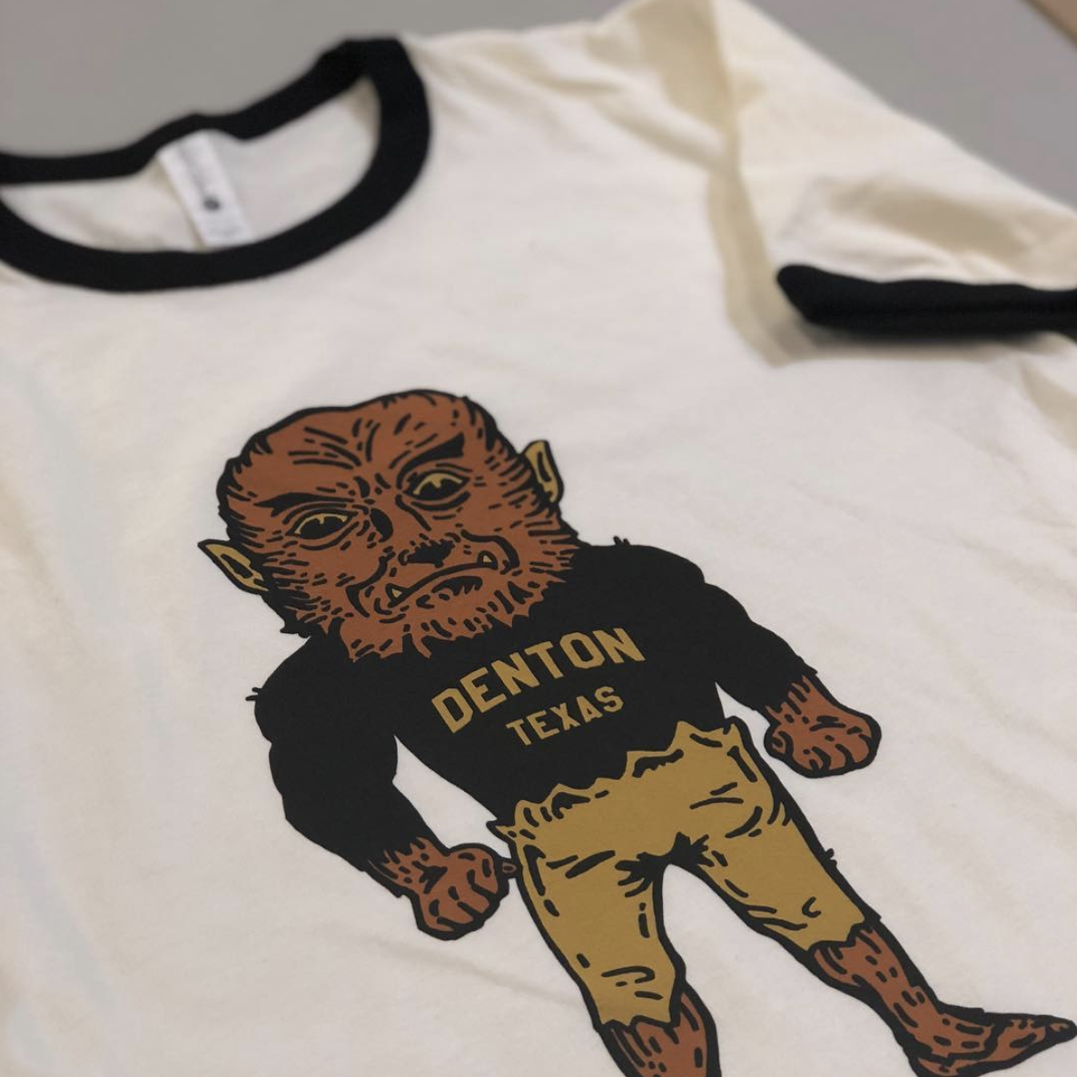 A white T-shirt with black collar and sleeve trim featuring a cartoon graphic of an ape in a black shirt with yellow text 'DENTON TEXAS' and tan pants.