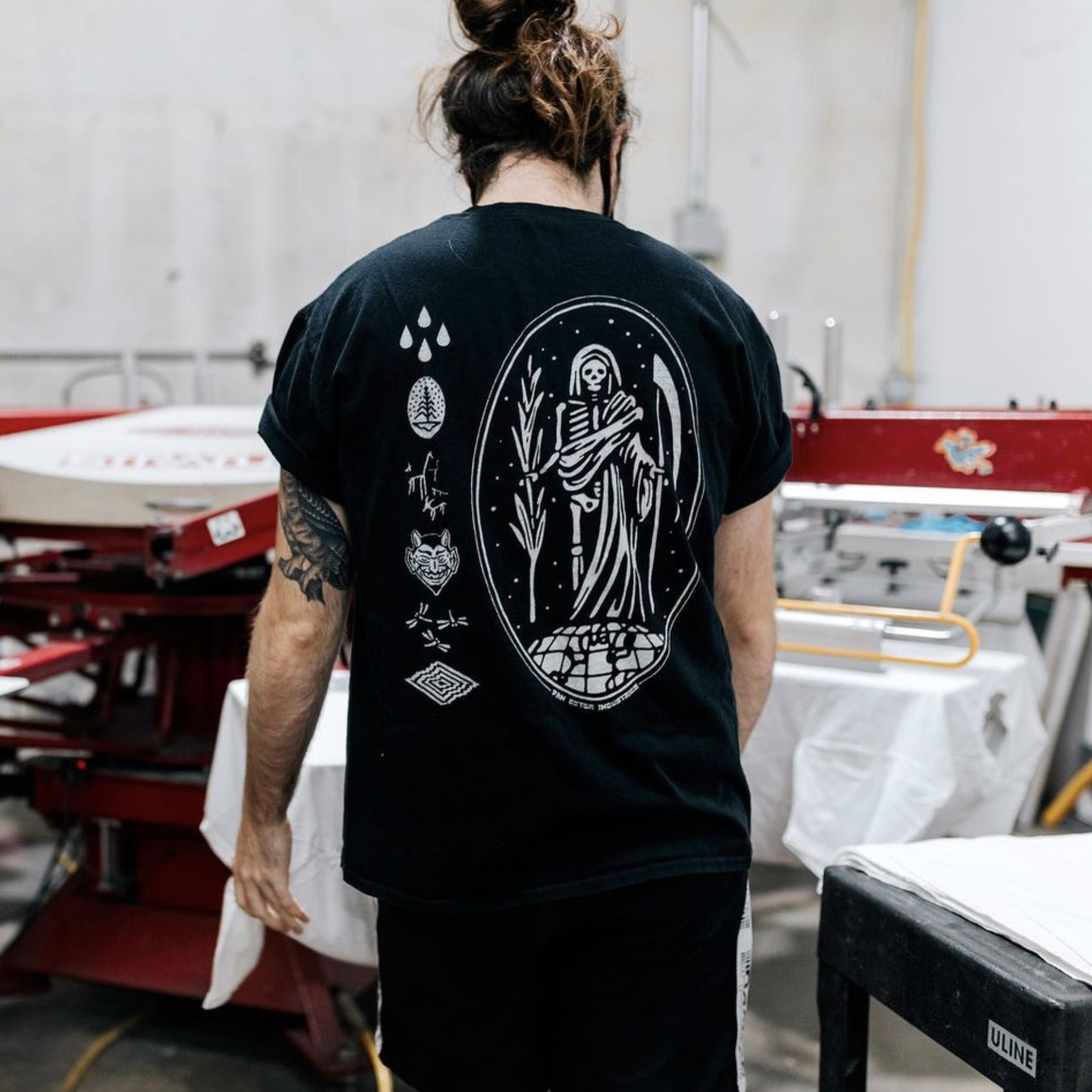A person with tattooed arms is wearing a black T-shirt with white graphic illustrations on the back. The design features a skeletal figure in robes, standing amid plants, with additional symbols like drops, a mask, and geometric shapes around it. The