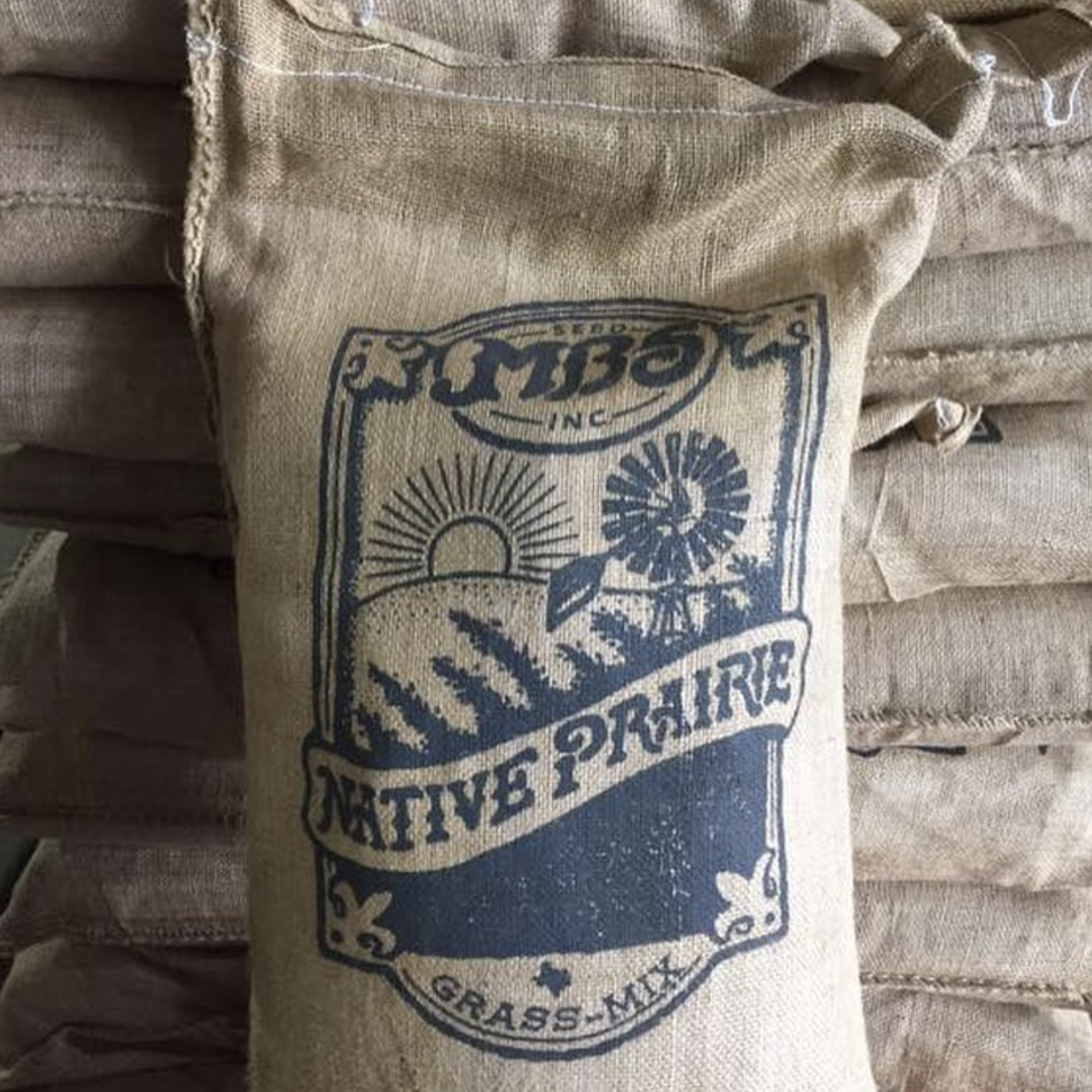A burlap sack with the branding of 'Revolution Inc. Active Nature' and the phrase 'Grass-Mix' written on it, featuring a sun, windmill, and plant illustrations.