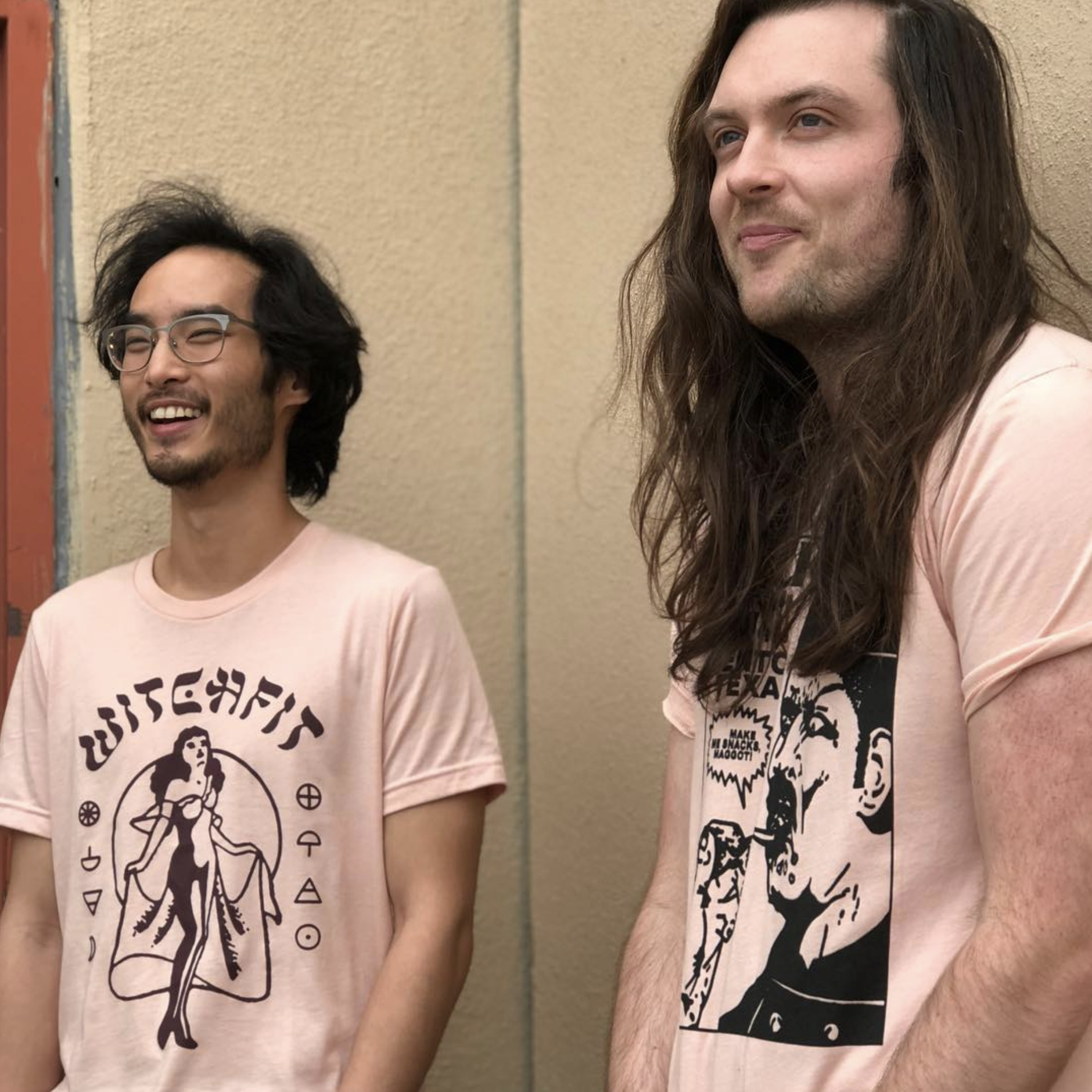 Two young men smiling, standing next to a beige wall. The man on the left has glasses, dark wavy hair, and wears a light pink T-shirt with a witchcraft-themed graphic. The man on the right has long brown hair, and wears a light pink T-shirt with a bl