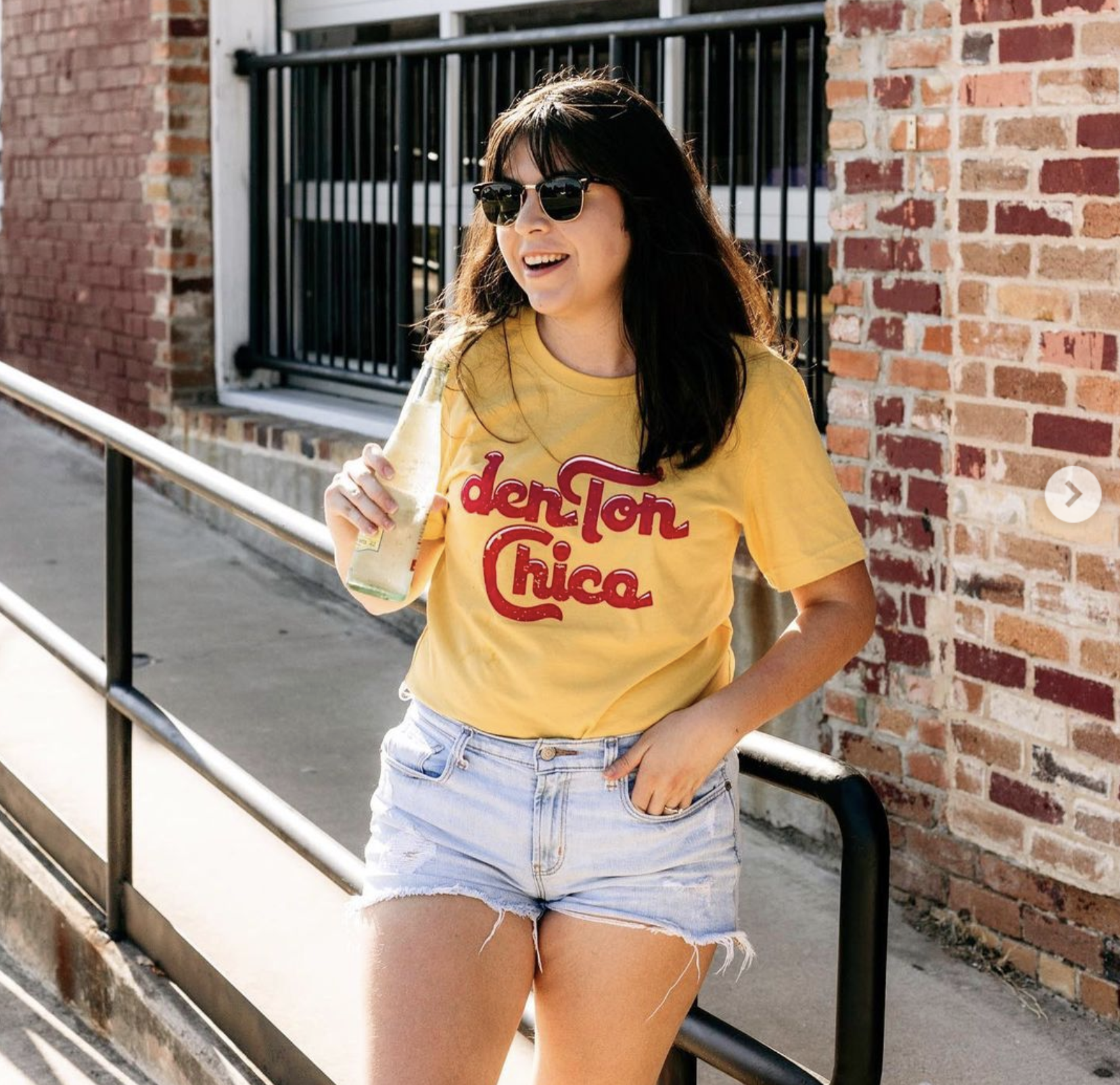 A young woman with dark hair, wearing sunglasses, a yellow T-shirt with red text, and denim shorts, stands outdoors beside a brick wall, holding a bottle in her right hand, smiling and looking to her left.