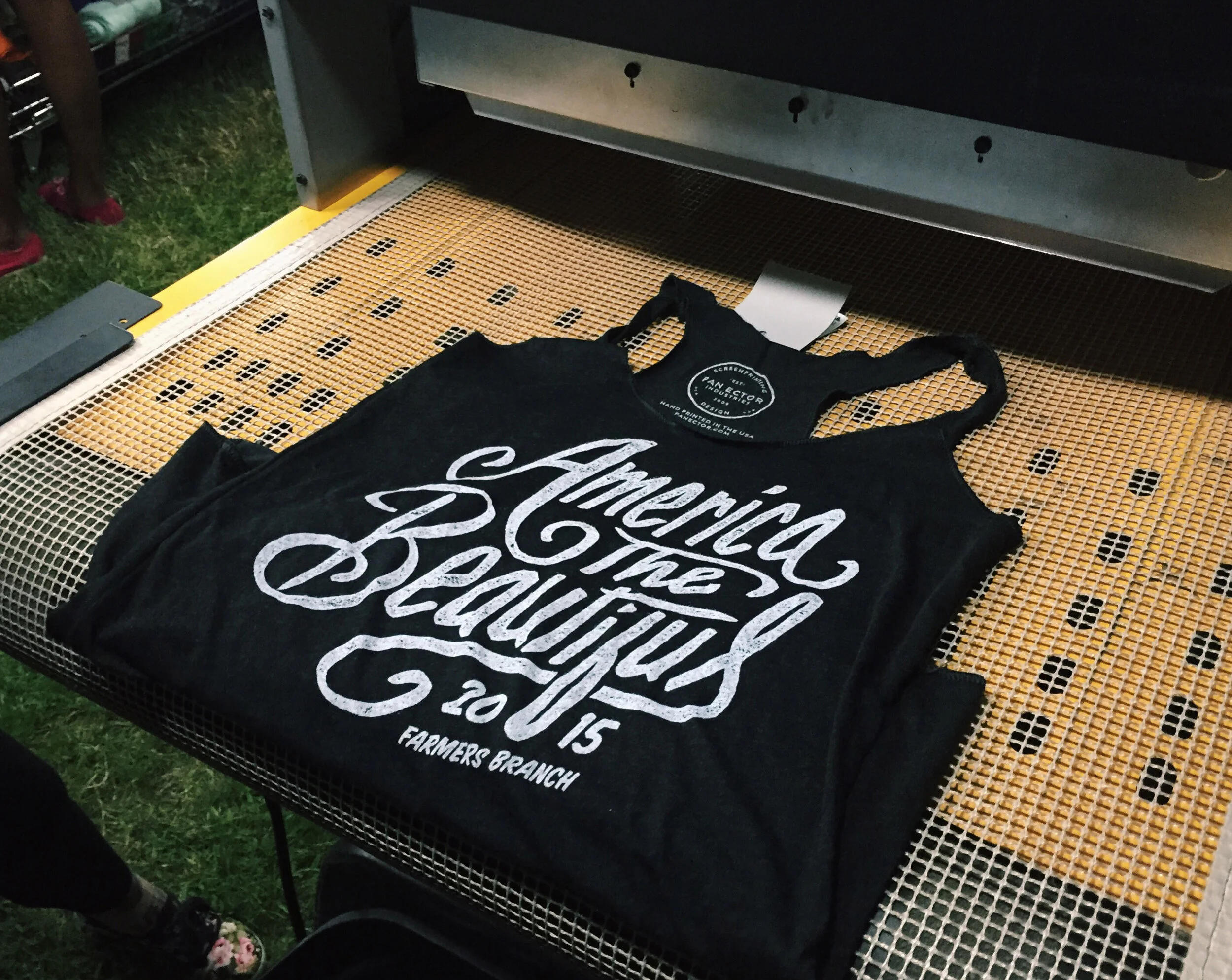 Black tank top with white text reading "American the Beautiful 2015 Farmers Branch" on a mesh surface.