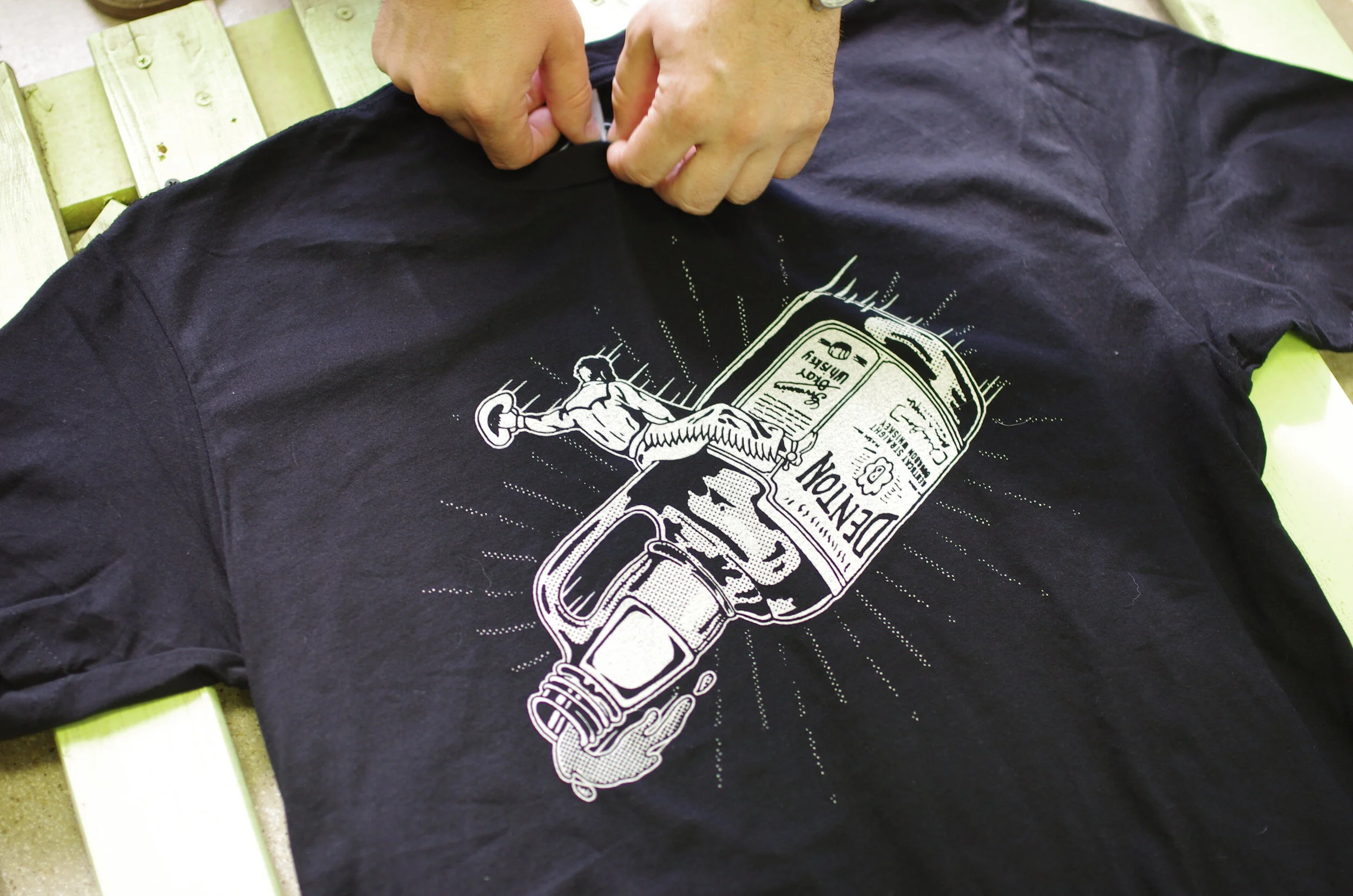 A person is heat pressing a white graphic design onto a black T-shirt. The design features a vintage bottle with a small muscular figure lifting a barbell, along with various labels and text.