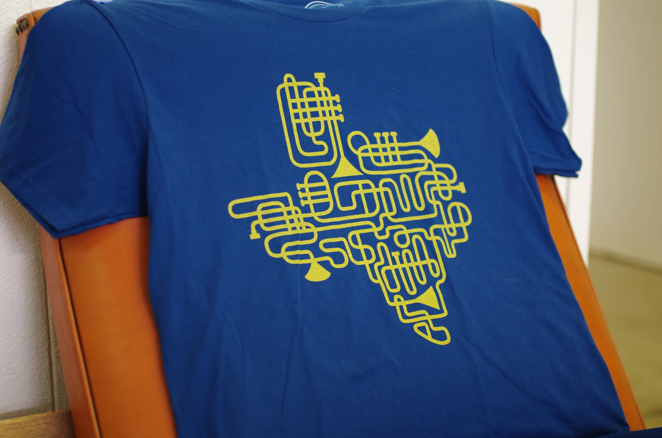 Blue T-shirt with yellow graphic of musical instruments like a trumpet, trombone, and saxophone