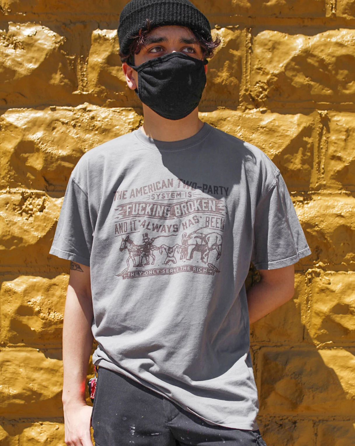 Young person wearing a black mask, gray t-shirt, black beanie, standing in front of a yellow brick wall.