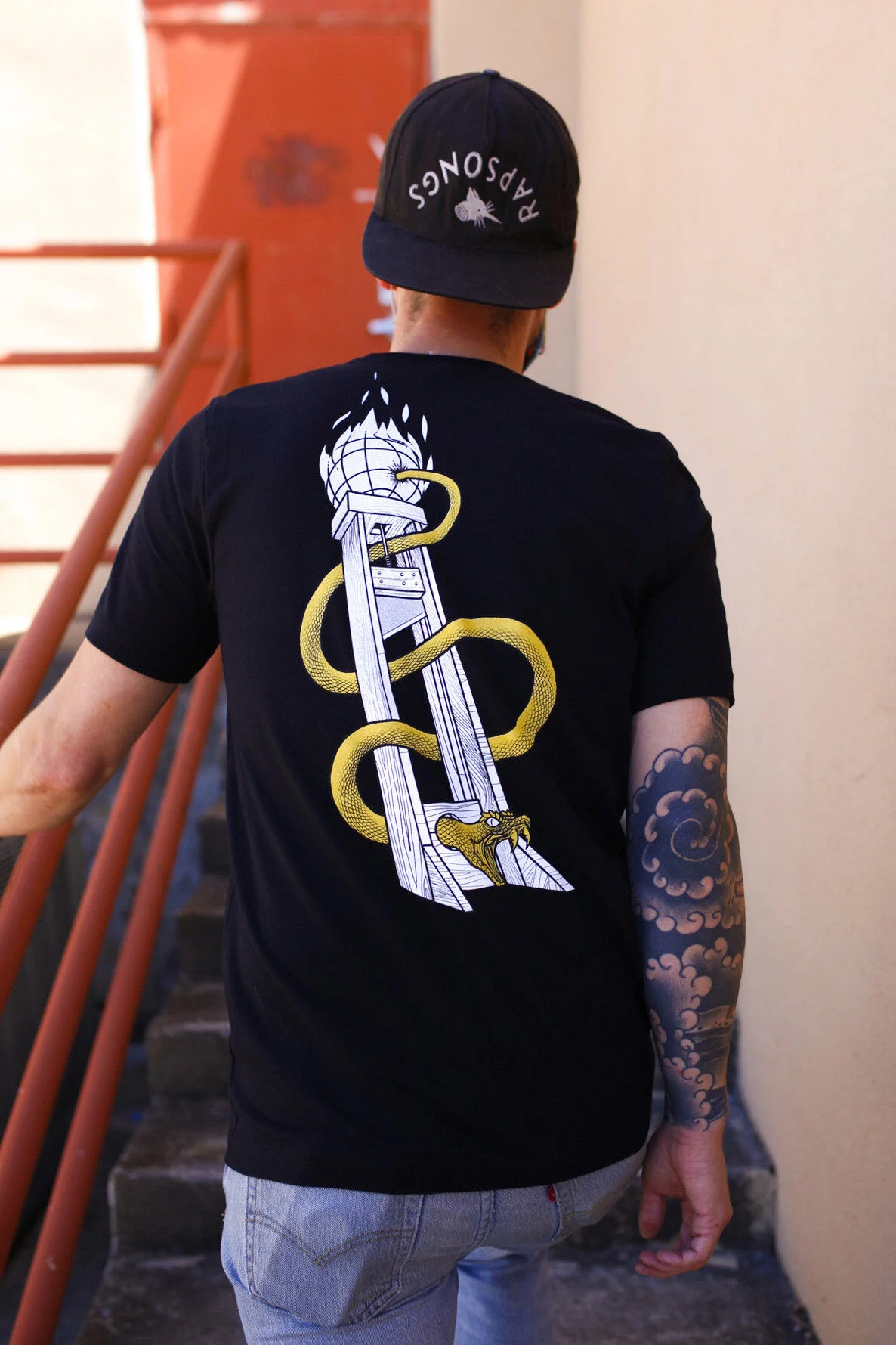 A person wearing a black baseball cap with white text and a graphic image, and a black T-shirt with a graphic design of a ladder, a snake, and a snake's head in yellow and white, walking down a staircase with an orange railing.