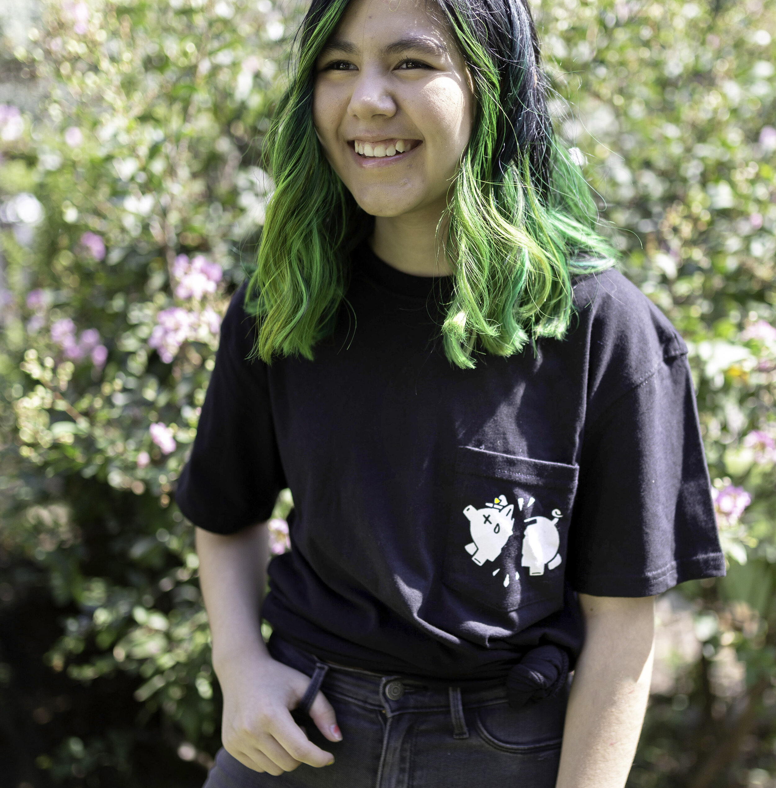 A young woman with shoulder-length green-haired hair, wearing a black t-shirt with a graphic of a cartoon animal, smiling outdoors with pink flowers and greenery in the background.
