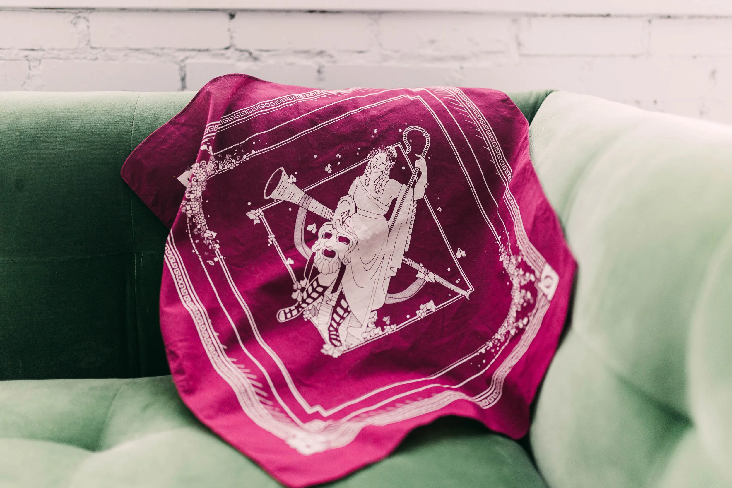 A pink bandana with white printed design resting on a green couch against a white brick wall.