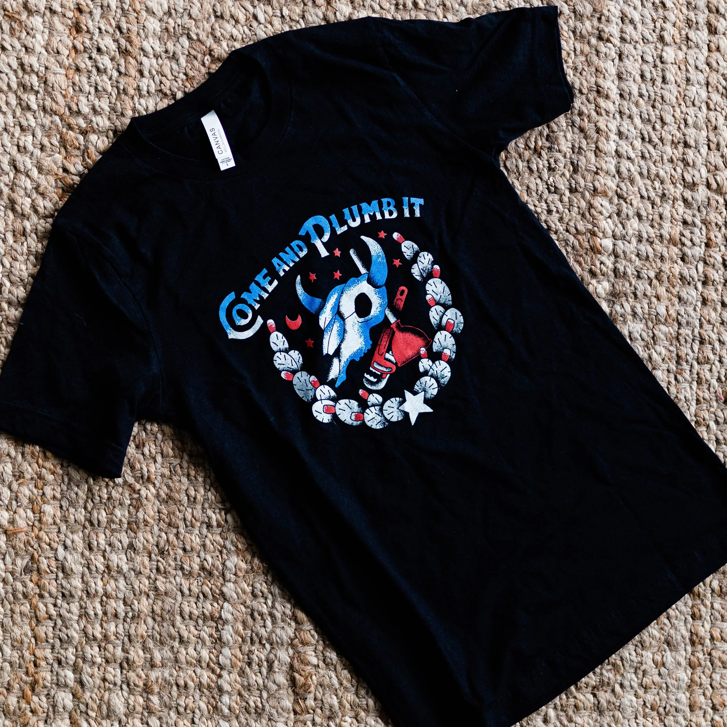 Black T-shirt with a graphic of a cow skull, a red cup, and the phrase 'Come and Plumb It' on a beige carpet.