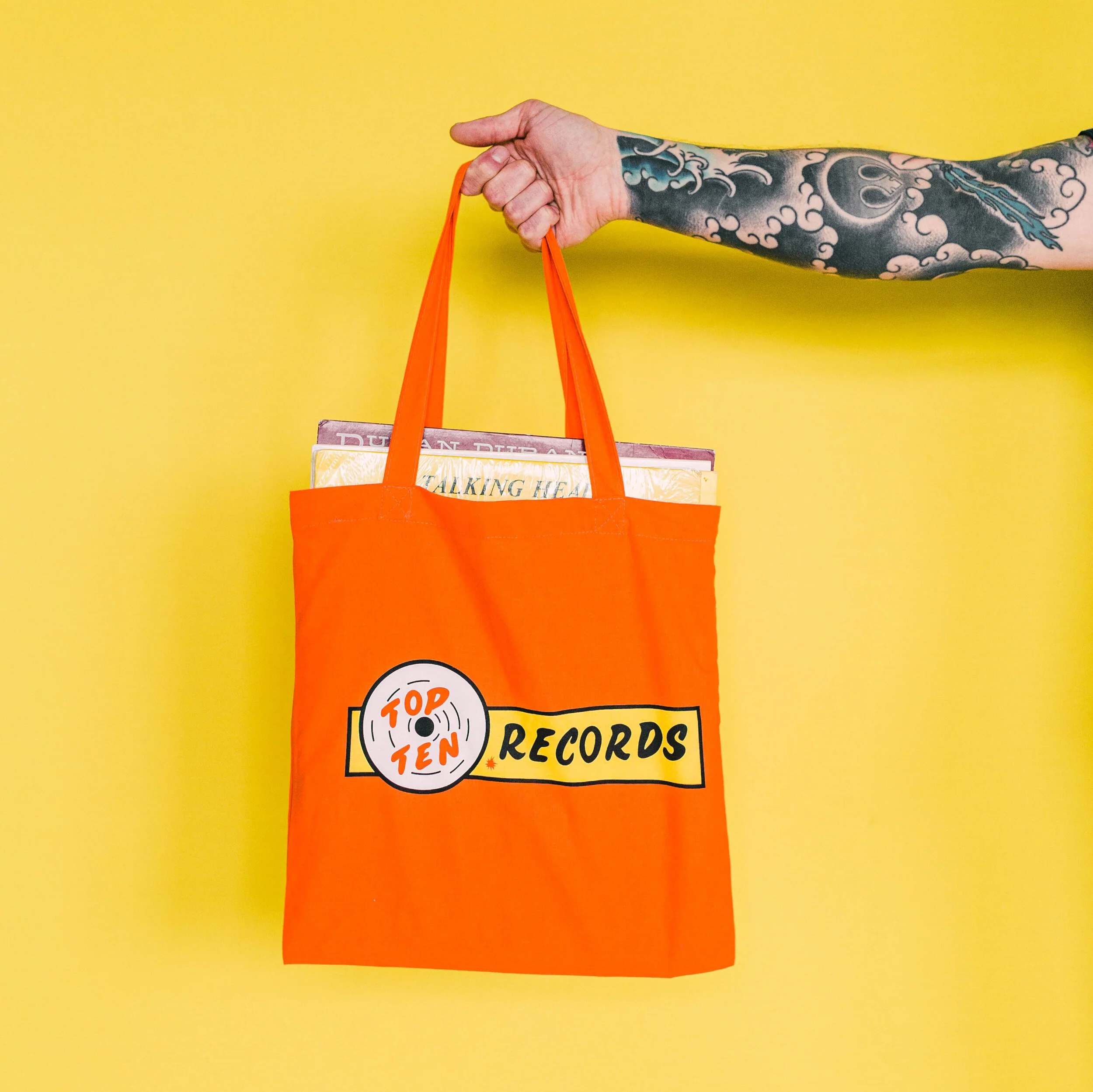 A person with a tattooed arm holding an orange tote bag with 'Top Ten Records' logo, against a yellow background.
