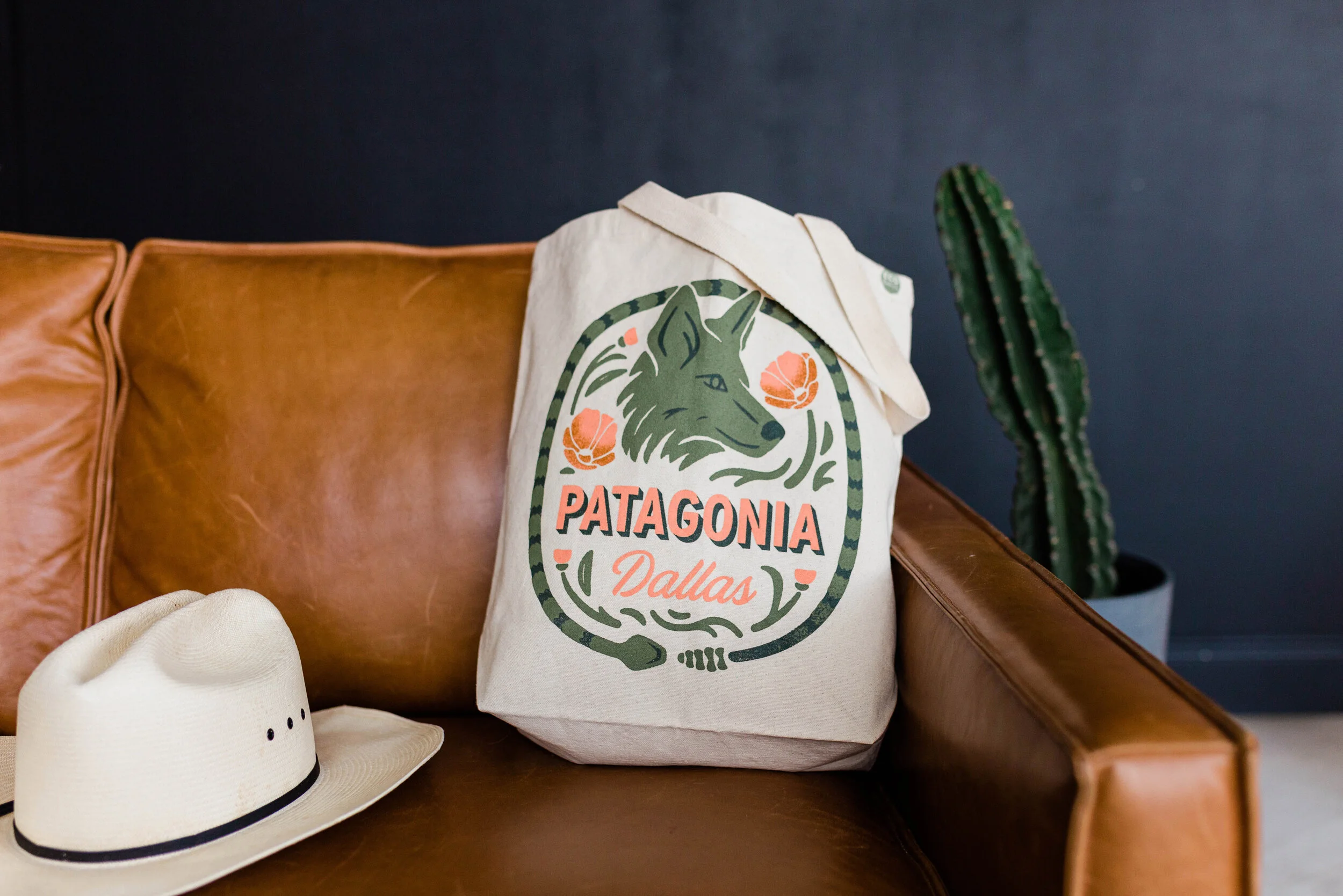 A tote bag with a Patagonia Dallas logo featuring a wolf's head and floral elements, placed on a brown leather couch, with a white hat and a tall potted cactus nearby.