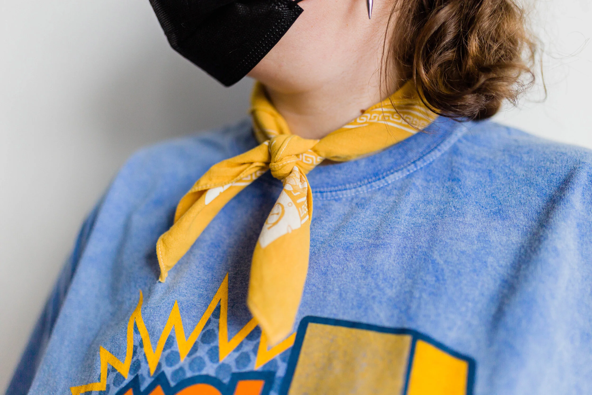 Close-up of a person wearing a black face mask, yellow neckerchief, blue sweatshirt, and a graphic T-shirt.