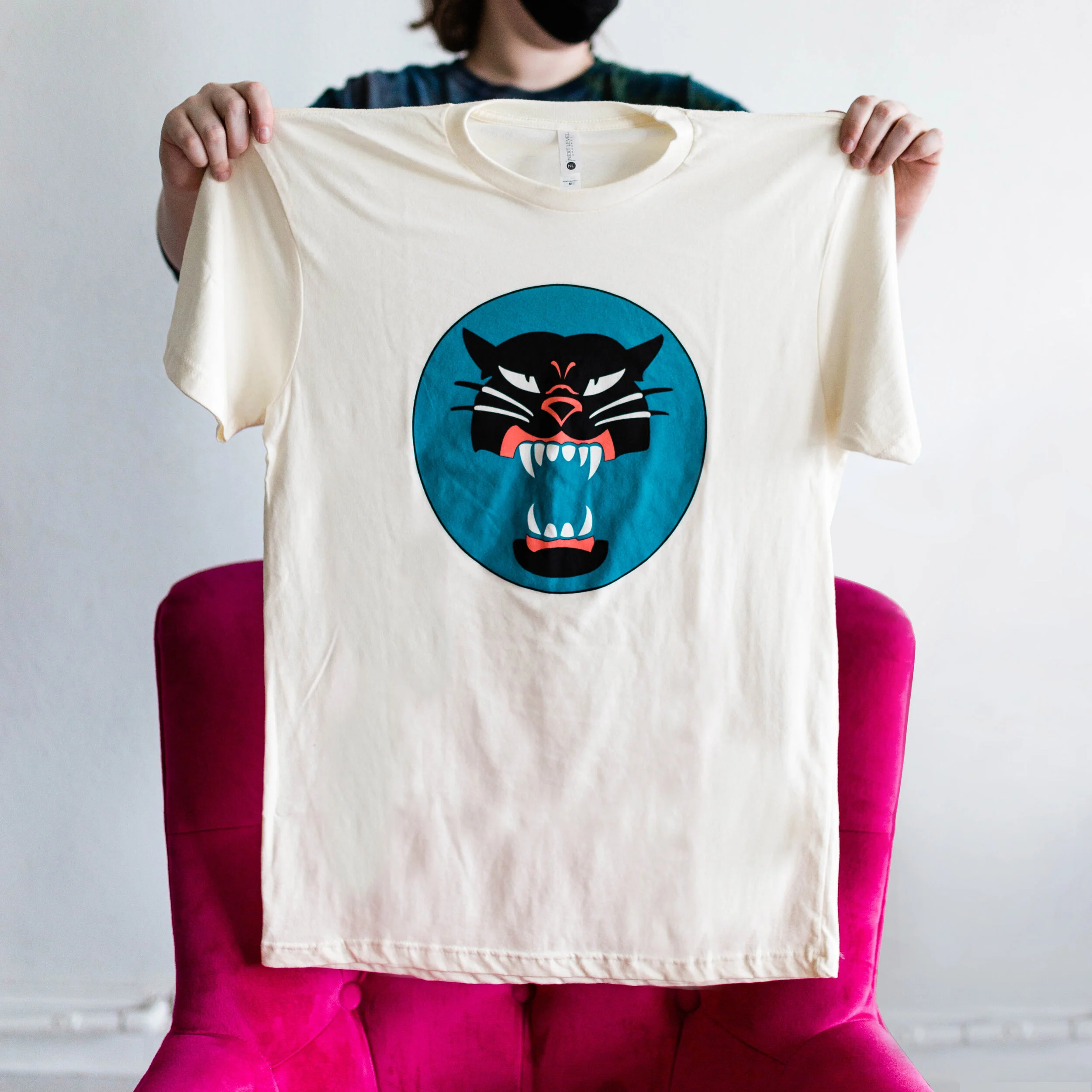 Person holding a cream-colored t-shirt with a large graphic of a black panther's head, open mouth showing teeth, inside a blue circle.