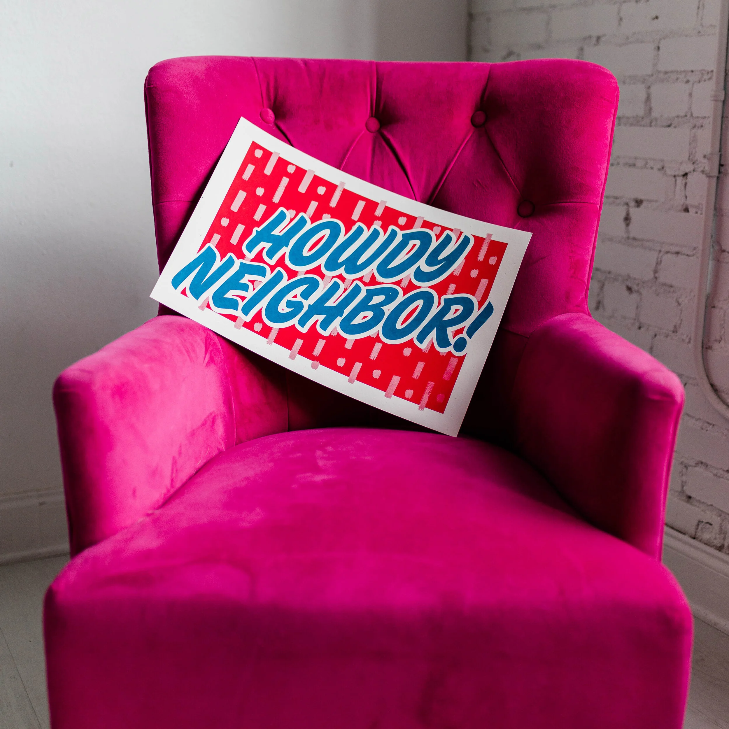 A bright pink velvet armchair with a sign that reads "Howdy Neighbor!" leaning against the backrest.