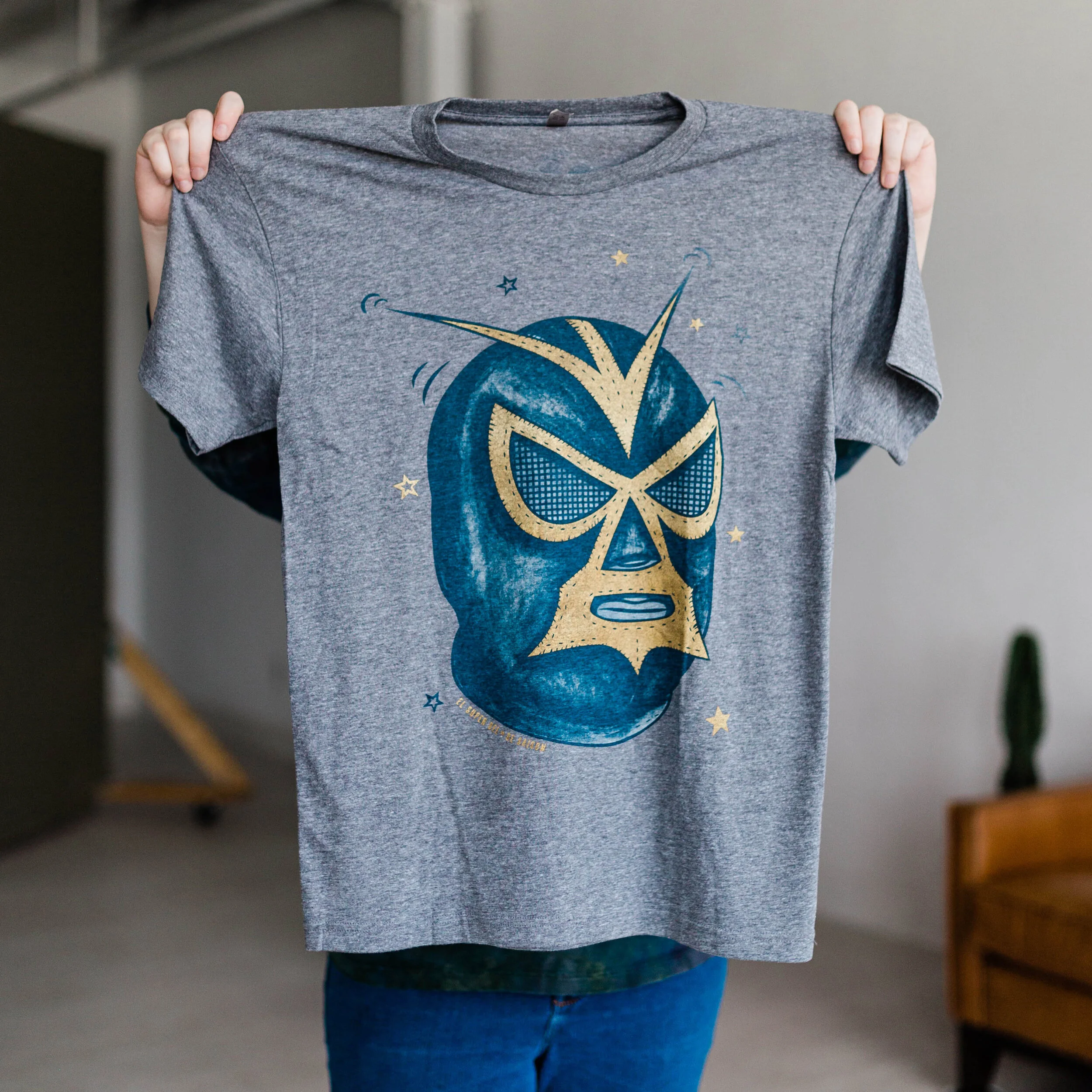 Person holding up a gray T-shirt with a blue and gold luchador mask design on it.