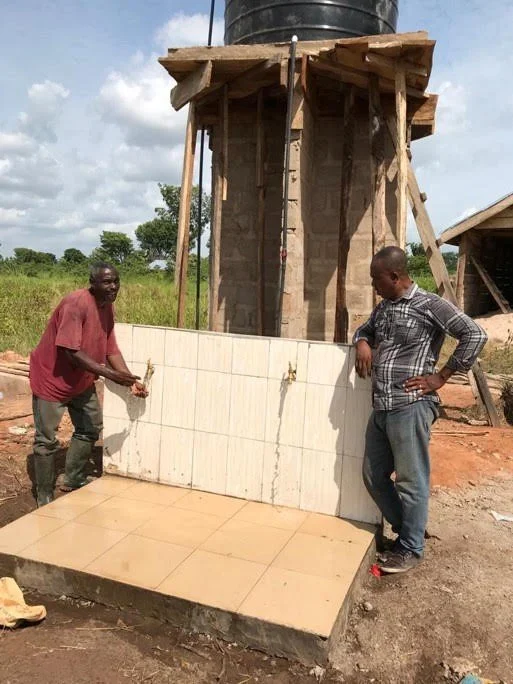 Two men stand next to a tiled wall and floor section under construction, with a water tank structure behind them in a rural setting.