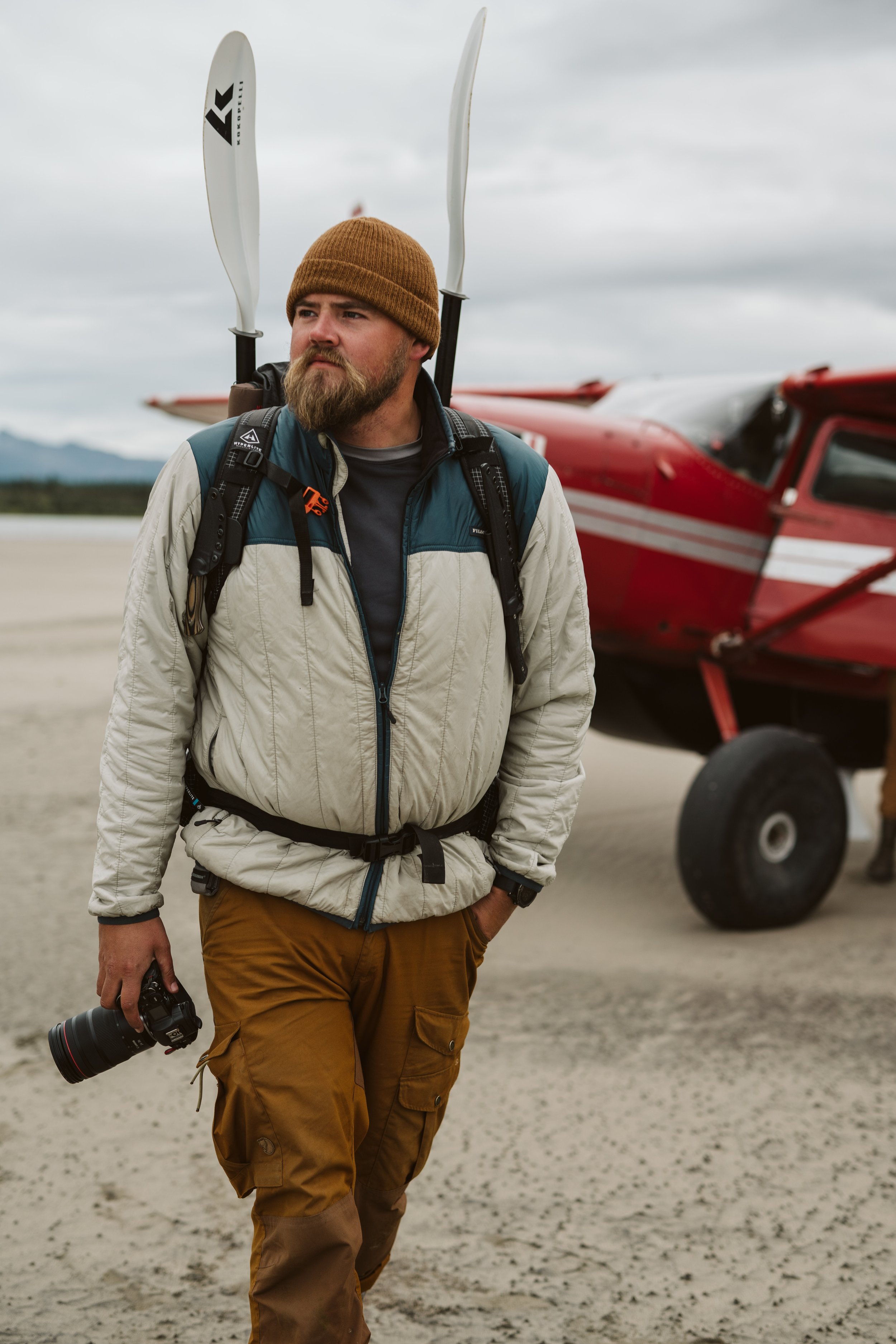 Very large man holding a camera and walking in front of a red single prop plane in Alaska