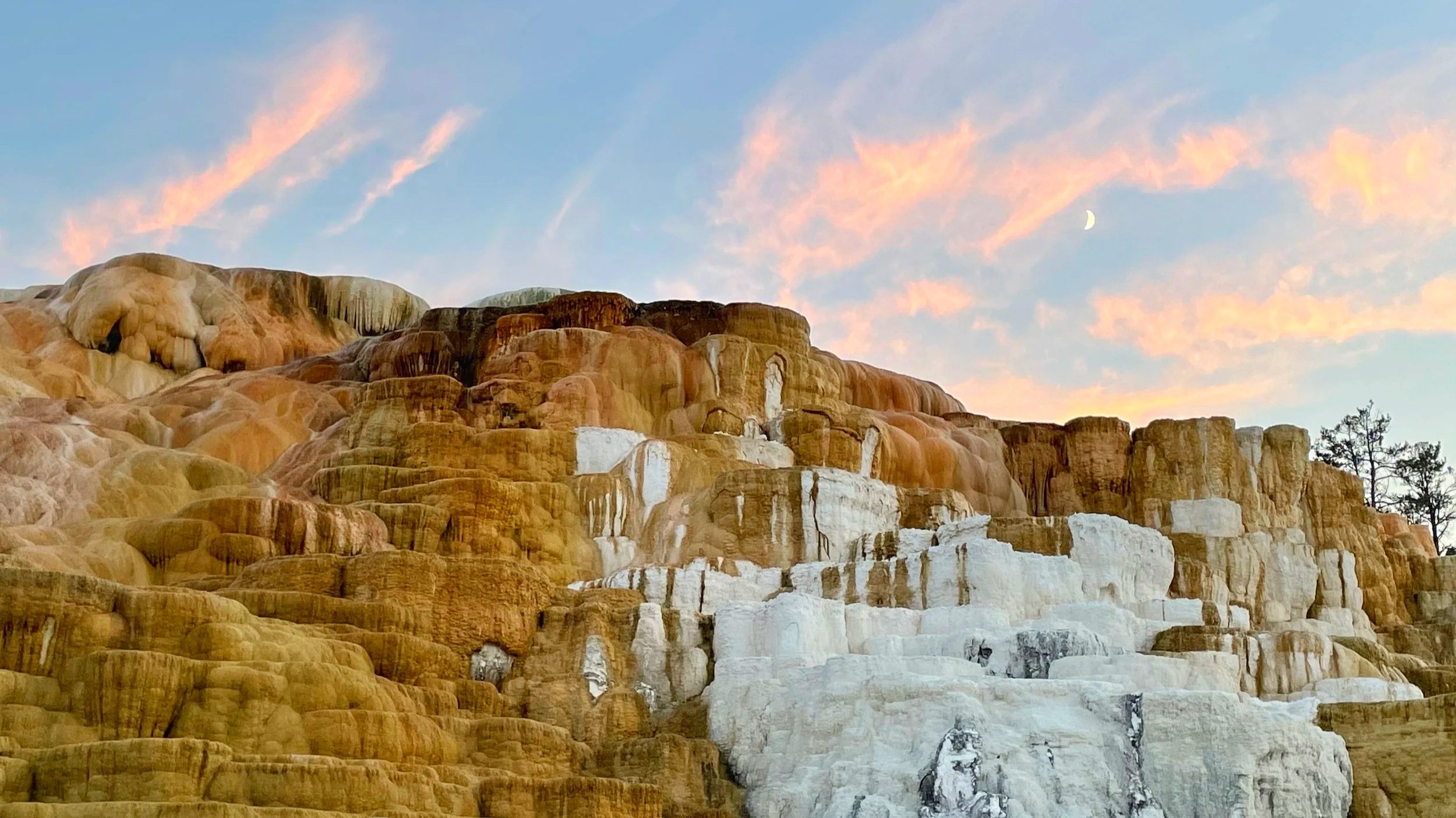 Mammoth Hot Springs in Yellowstone are orange with a sunset sky and moon above