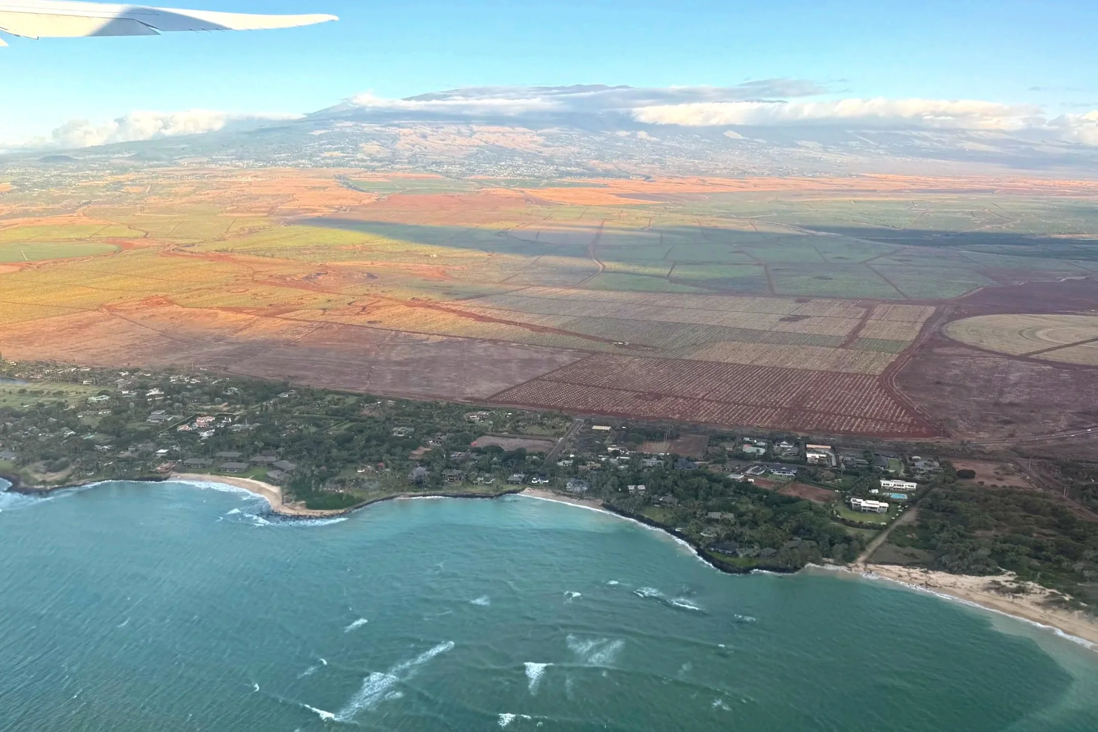 An airplane flies over the ocean and fields in Hawaii.