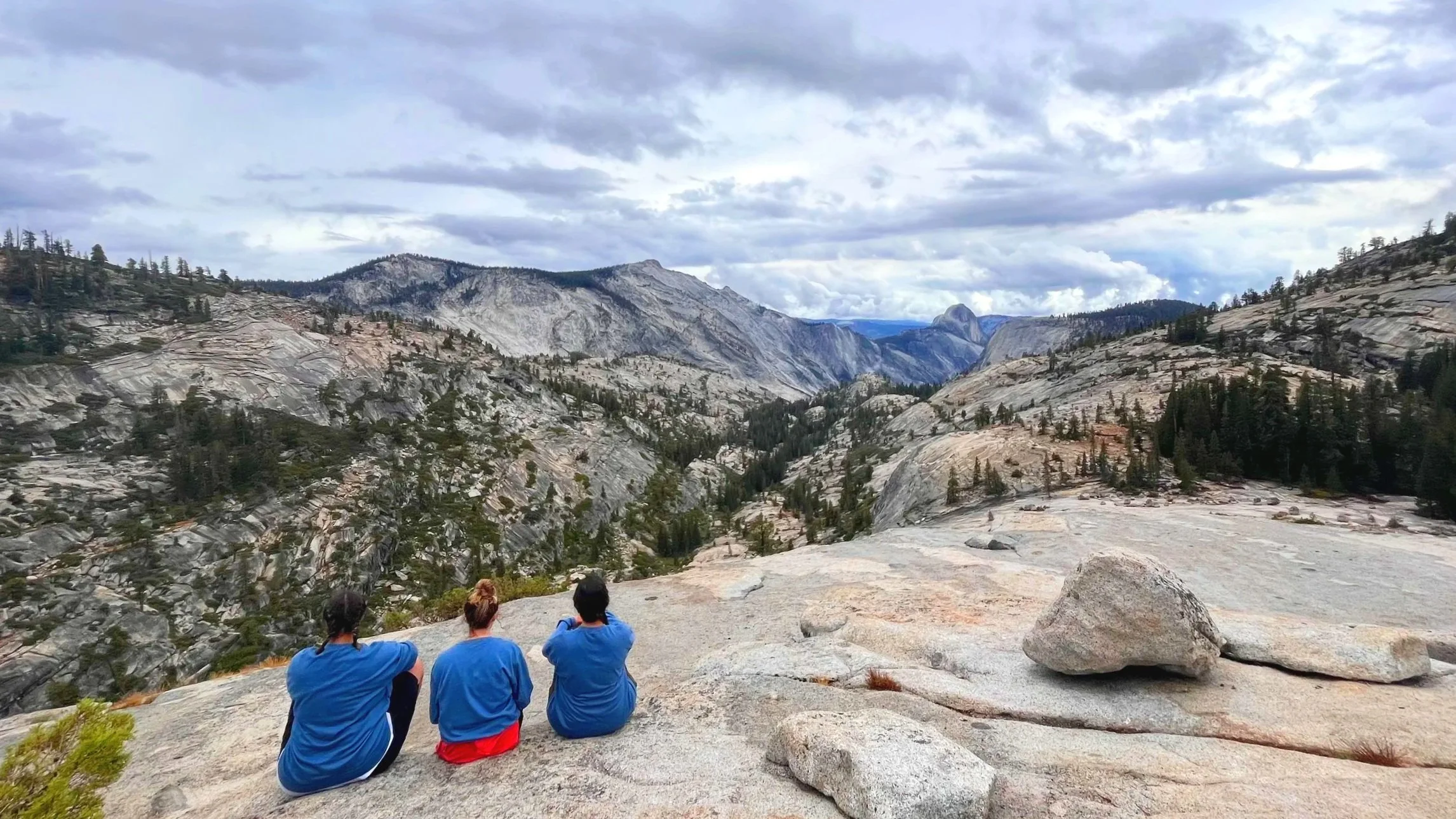 Three matching women sit on a granite cliff overlooking sweeping views of mountains in the distance.