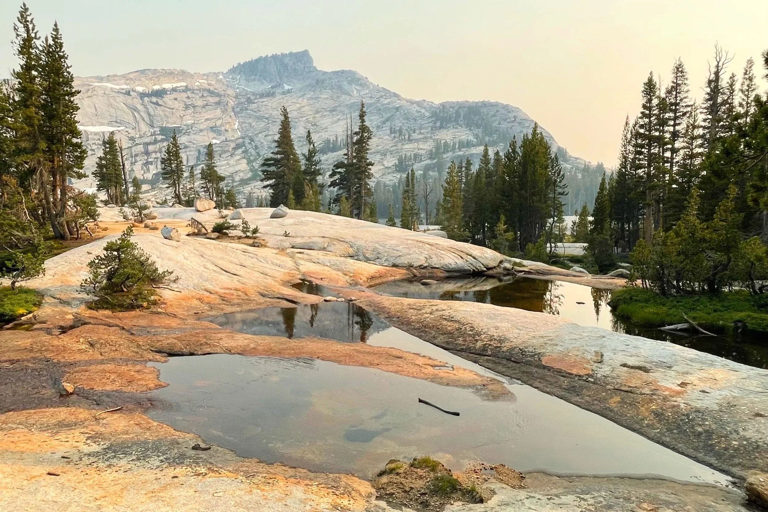 Pools of water in the forefront of the picture are surrounded by pine trees and granite mountains are seen in the background.
