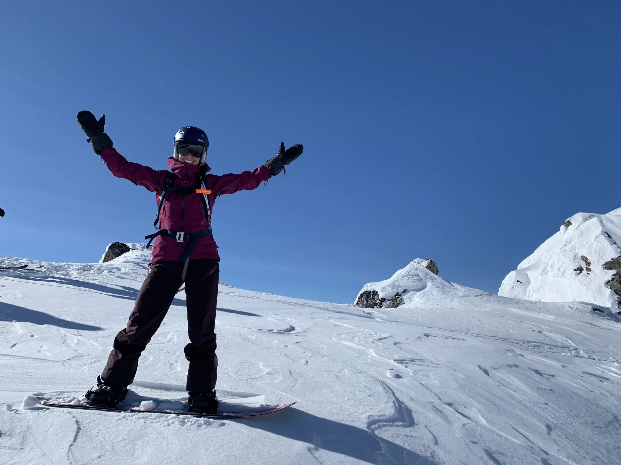 Person in ski gear standing on snow with arms outstretched on a mountain slope under blue sky