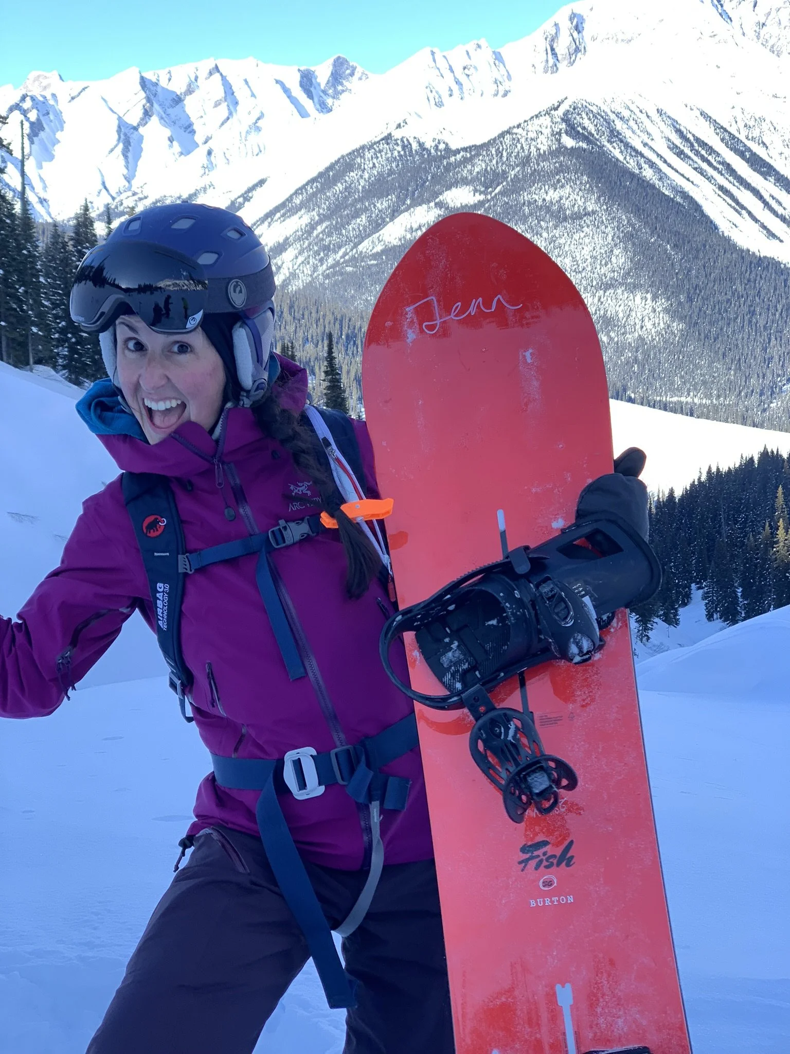 A woman wearing a purple ski jacket, black helmet, and black gloves holding a red snowboard with bindings, smiling in a snowy mountain landscape with evergreen trees and snow-covered peaks in the background.