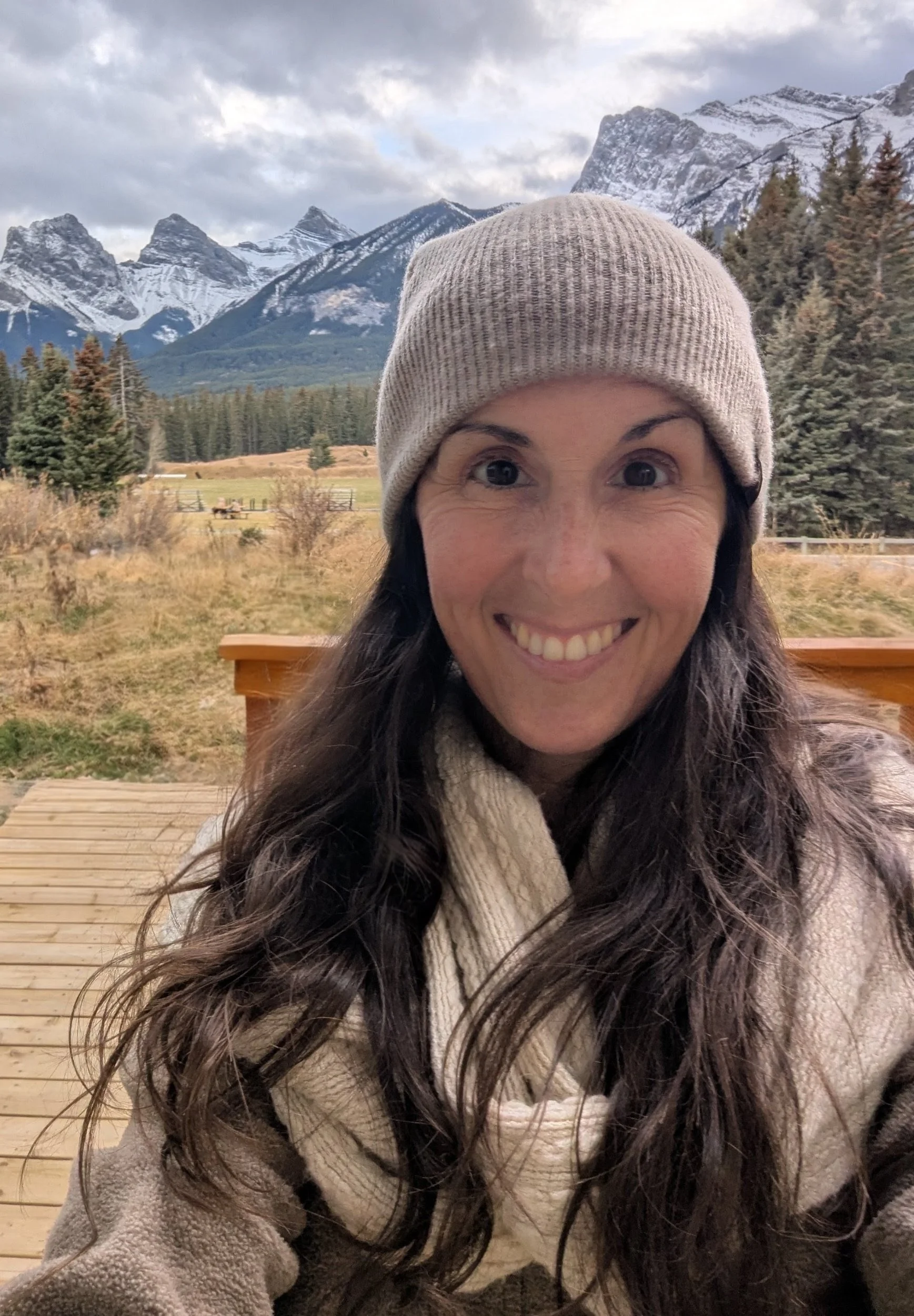 A woman smiling outdoors with snow-capped mountains and pine trees in the background, wearing a beige knit beanie and a scarf.