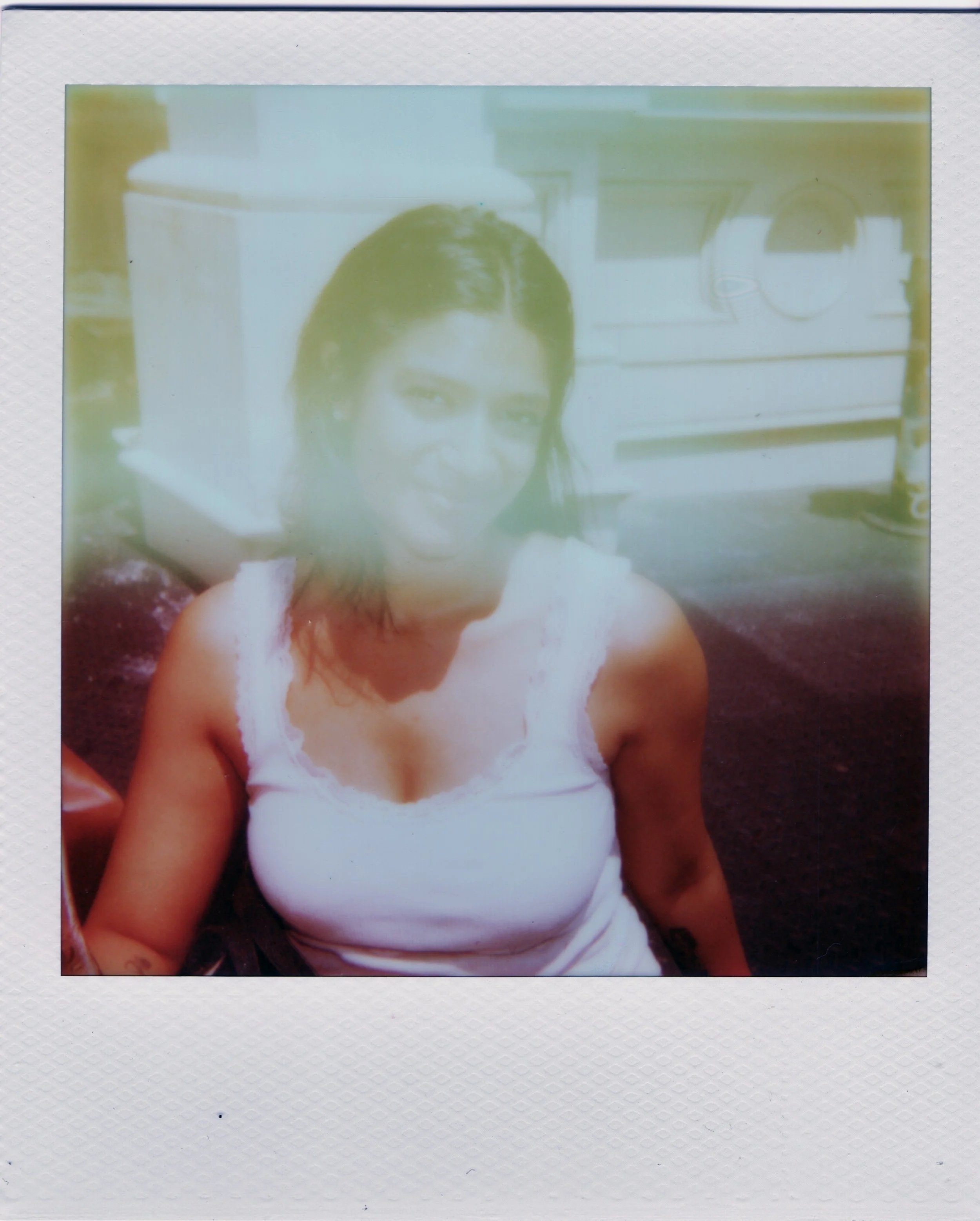 A woman with dark hair and glasses taking a selfie in a room with kitchen shelves in the background. She is wearing a white top and is leaning on a surface.