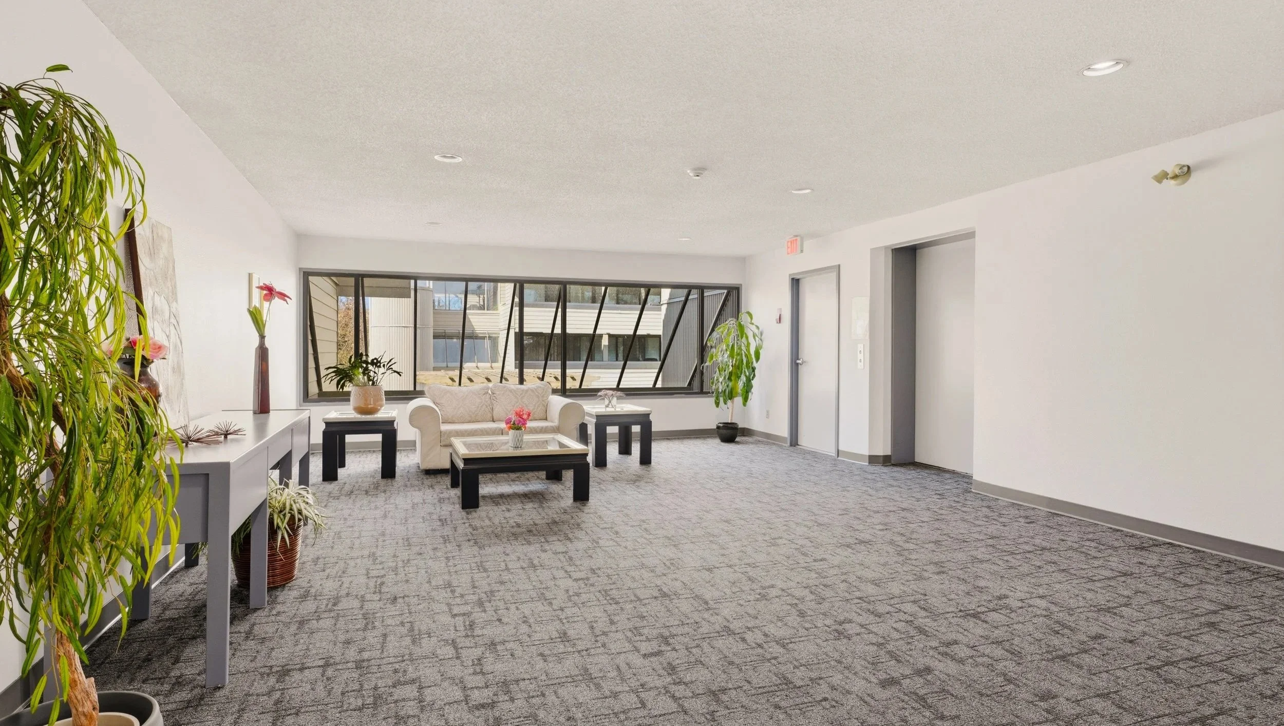 Interior of a lobby or waiting area with a sofa, coffee table, side tables, potted plants, and large window.