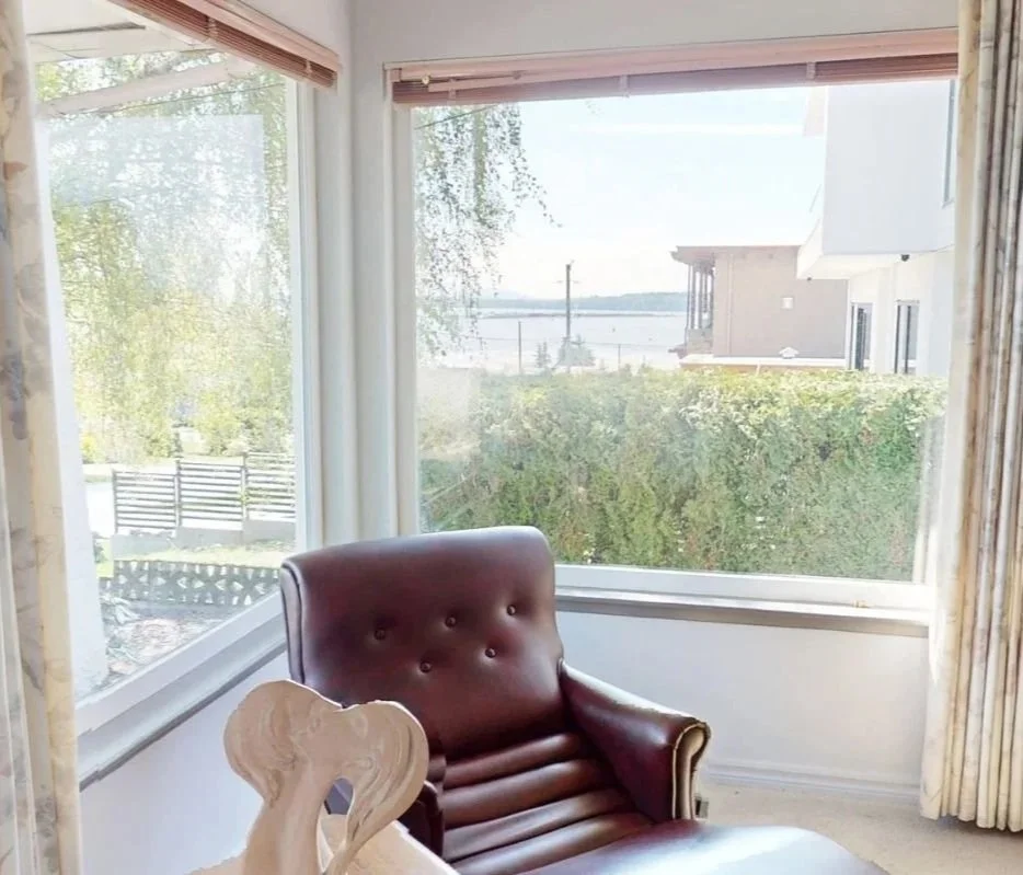 A corner of a room with large windows showing a view of the outdoors with trees, a hedge, and water in the background. Inside, there is a leather armchair and a decorative piece resembling a white heart-shaped sculpture.