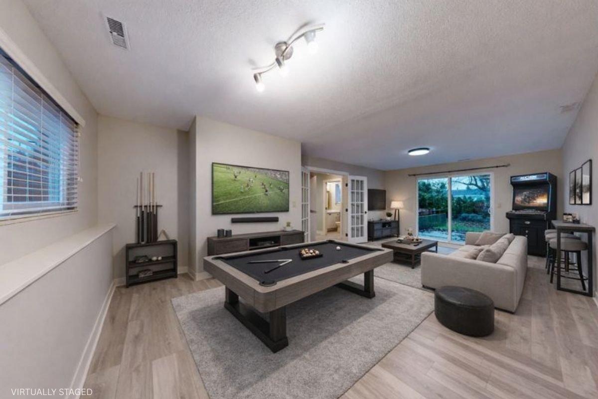 Living room with a pool table, beige sofa, and a large window showing a backyard with trees.