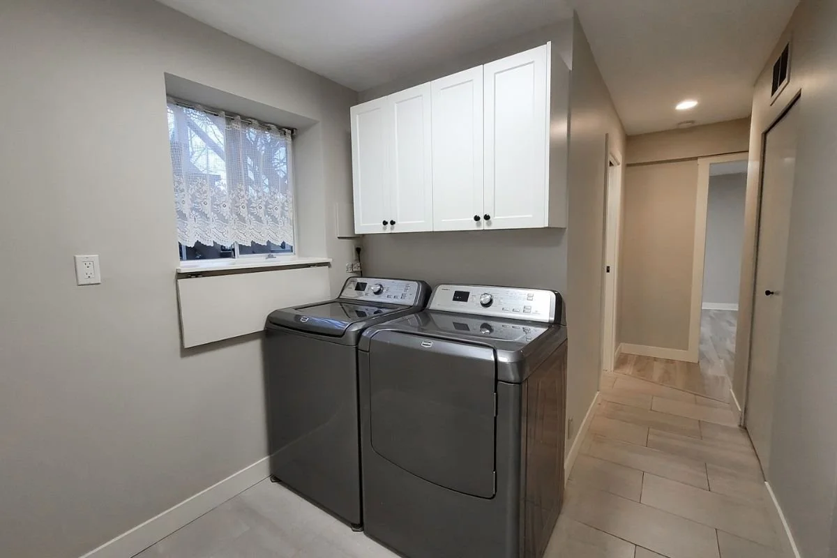 A laundry room with a washing machine and dryer, white cabinets above, a small window with lace curtains, and a hallway leading to other rooms.