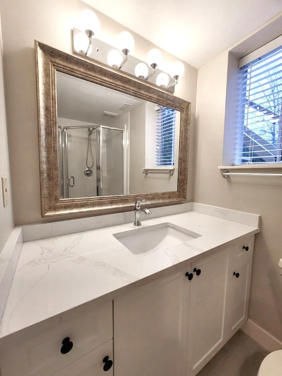 Bathroom vanity with white marble countertop, a rectangular undermount sink, silver faucet, large mirror, four globe lights above, two windows with blinds, and a shower with a glass door in the background.