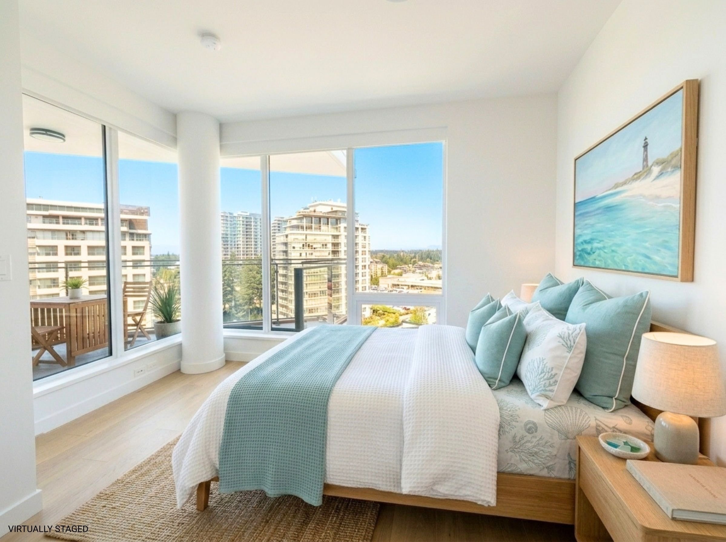 A bedroom with large windows showing a view of high-rise buildings and green trees outside. The room has a bed with a white and blue bedspread, multiple pillows, a wooden side table with a lamp and decorative bowl, and a painting of a lighthouse on t