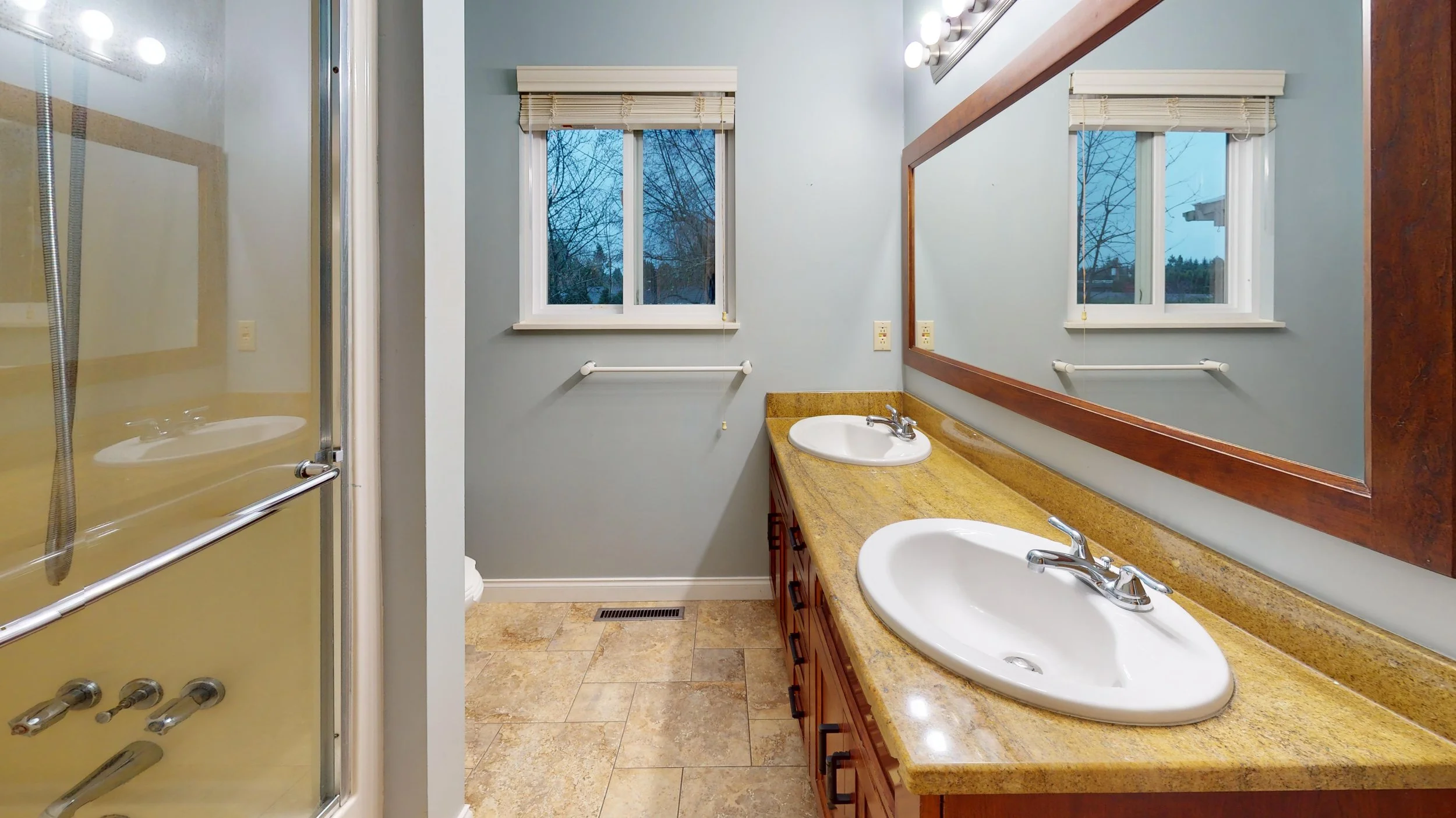 Bathroom with a double sink vanity with a granite countertop, a large mirror, a window showing an outdoor view, and a walk-in shower with a glass door.
