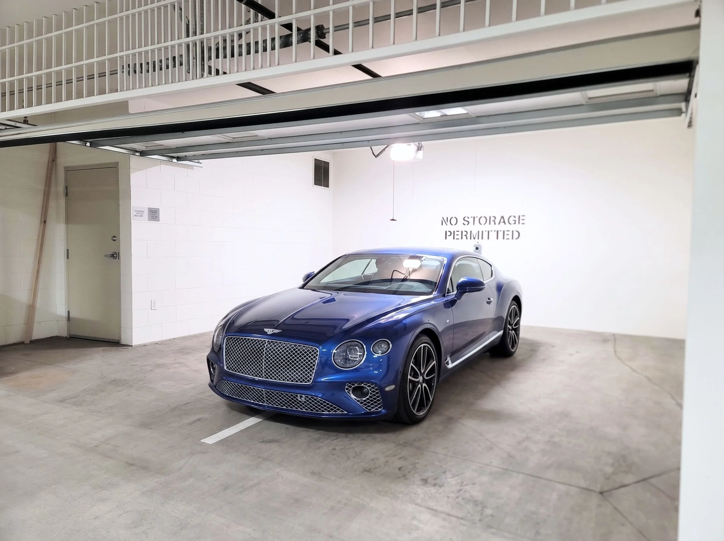 A blue luxury sports car parked in an underground parking garage with a sign on the wall that reads 'No Storage Permitted.'