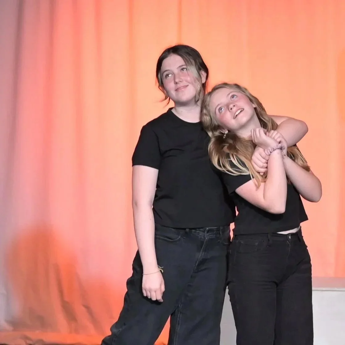 Two girls wearing black t-shirts, one hugging the other, standing on stage with a peach-colored curtain backdrop.