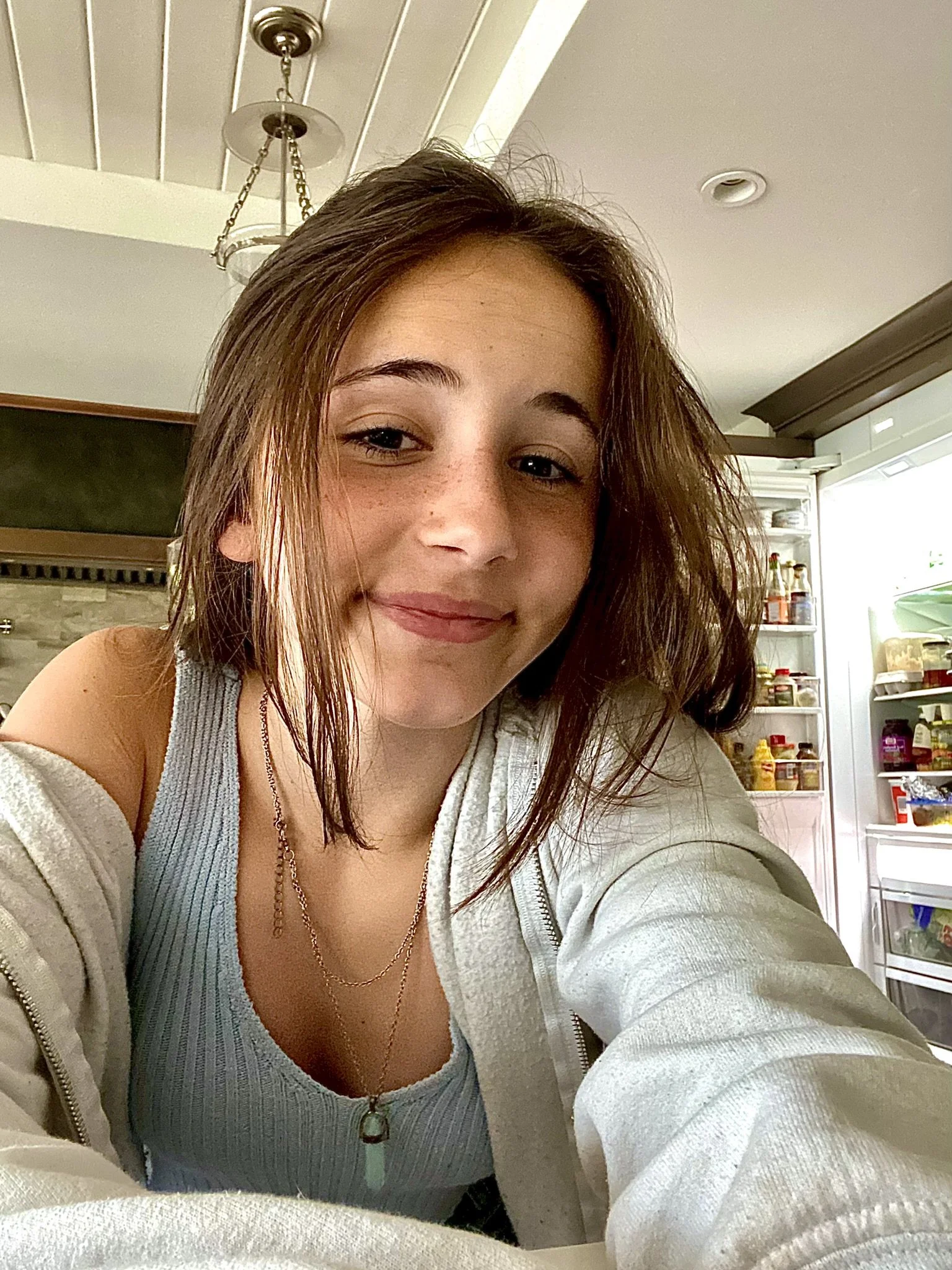 Close-up of a young woman smiling indoors with a refrigerator open behind her, showing various food and condiment bottles.