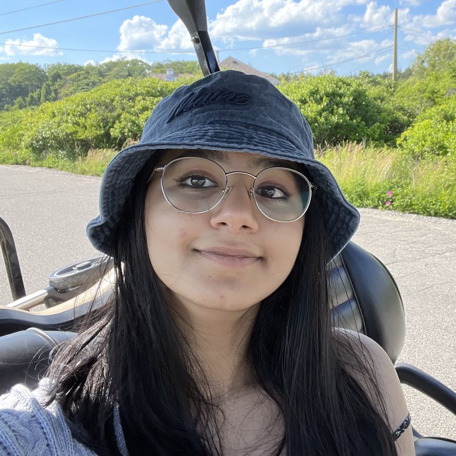 Young woman with long black hair, wearing a navy blue bucket hat, glasses, smiling, sitting on a motorcycle, outdoors on a sunny day with bushes and trees in the background.