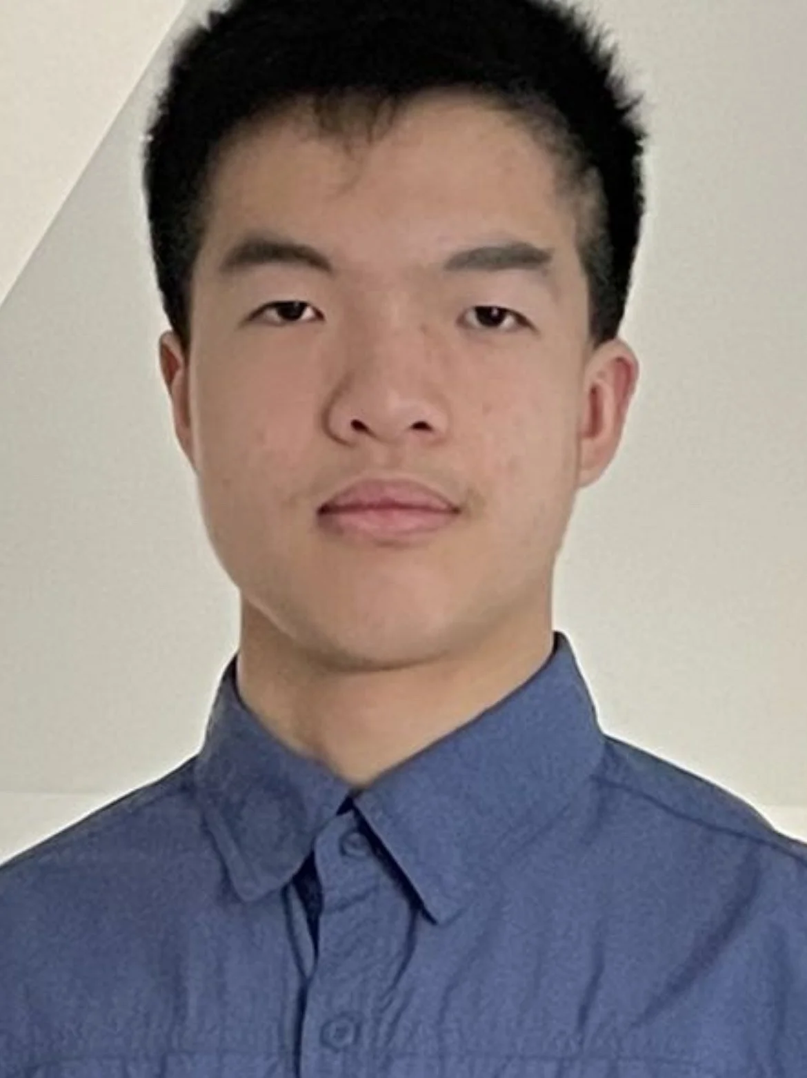 A young man with short black hair and light skin, wearing a blue button-up shirt, is looking at the camera with a neutral expression. The background is plain and off-white.