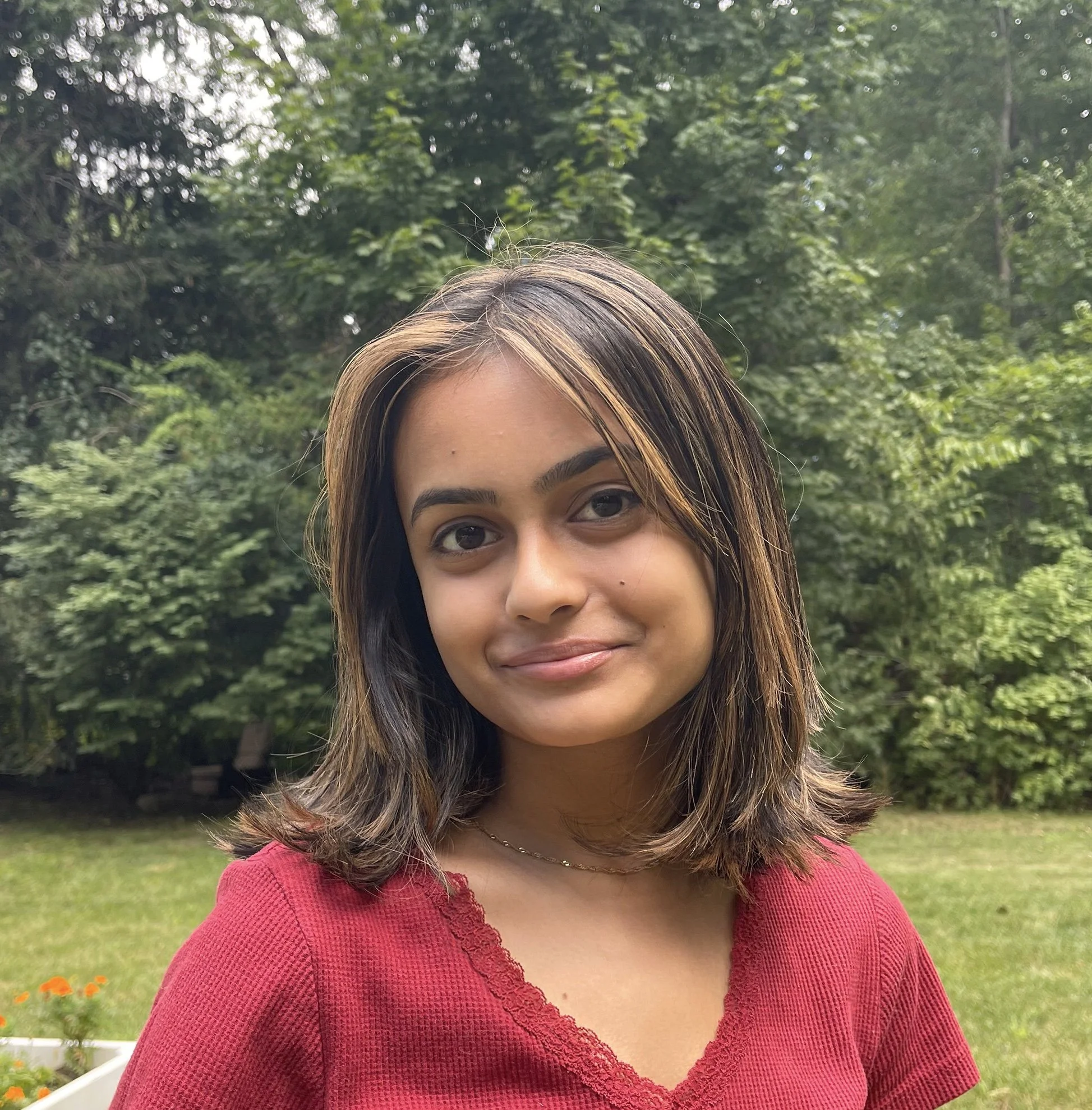 Young woman with shoulder-length brown hair and a red top, standing outdoors in front of green trees and grass.