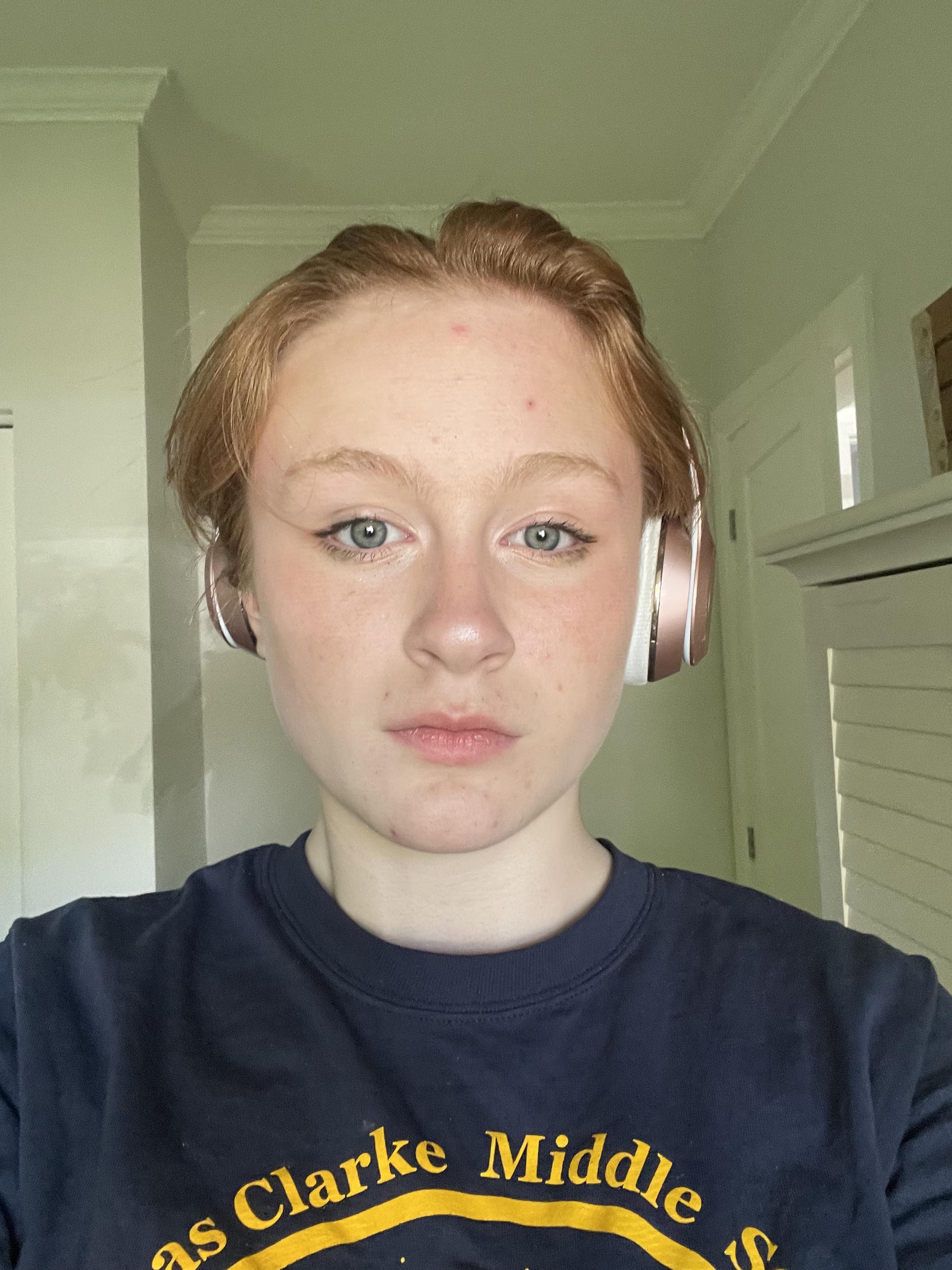 A young person with red hair and blue eyes wearing a black T-shirt with yellow text and large over-ear headphones, standing in a room with light green walls and white trim.