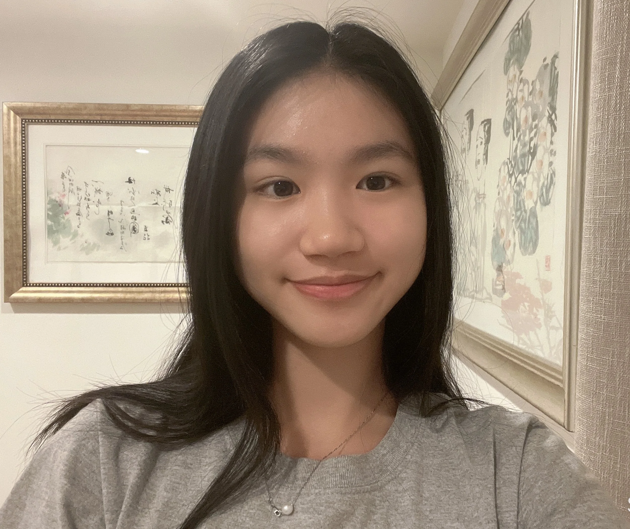 A young woman with long black hair taking a selfie indoors, smiling gently, wearing a gray sweatshirt and a pearl necklace, with Asian-style artwork in the background.