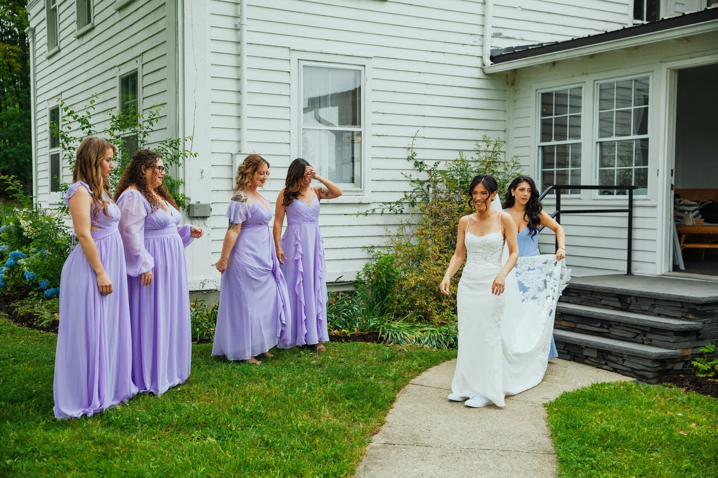A wedding party outdoors near a white house. The bride in a white dress is smiling and walking towards bridesmaids dressed in lavender gowns, who are standing on the grass and sidewalk.