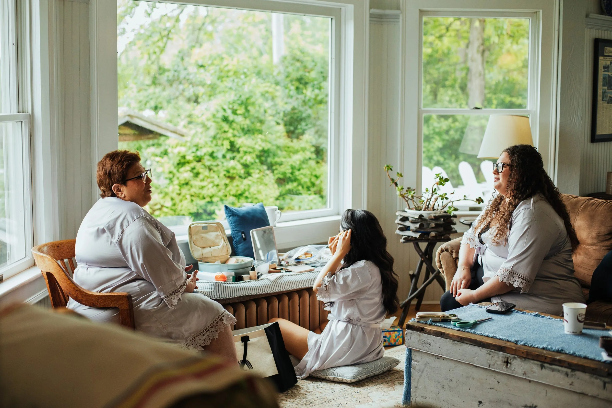 Three women in a cozy room with large windows overlooking green trees, engaged in conversation. One woman is seated on a chair, another on the floor with a pillow, and the third on a sofa. Various makeup and personal items are on the table.