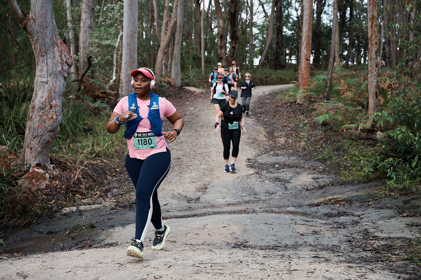 Group of people running on a forest trail during a race, with a woman in front wearing a pink shirt and blue vest, and others in the background following along the dirt path surrounded by tall trees and green foliage.