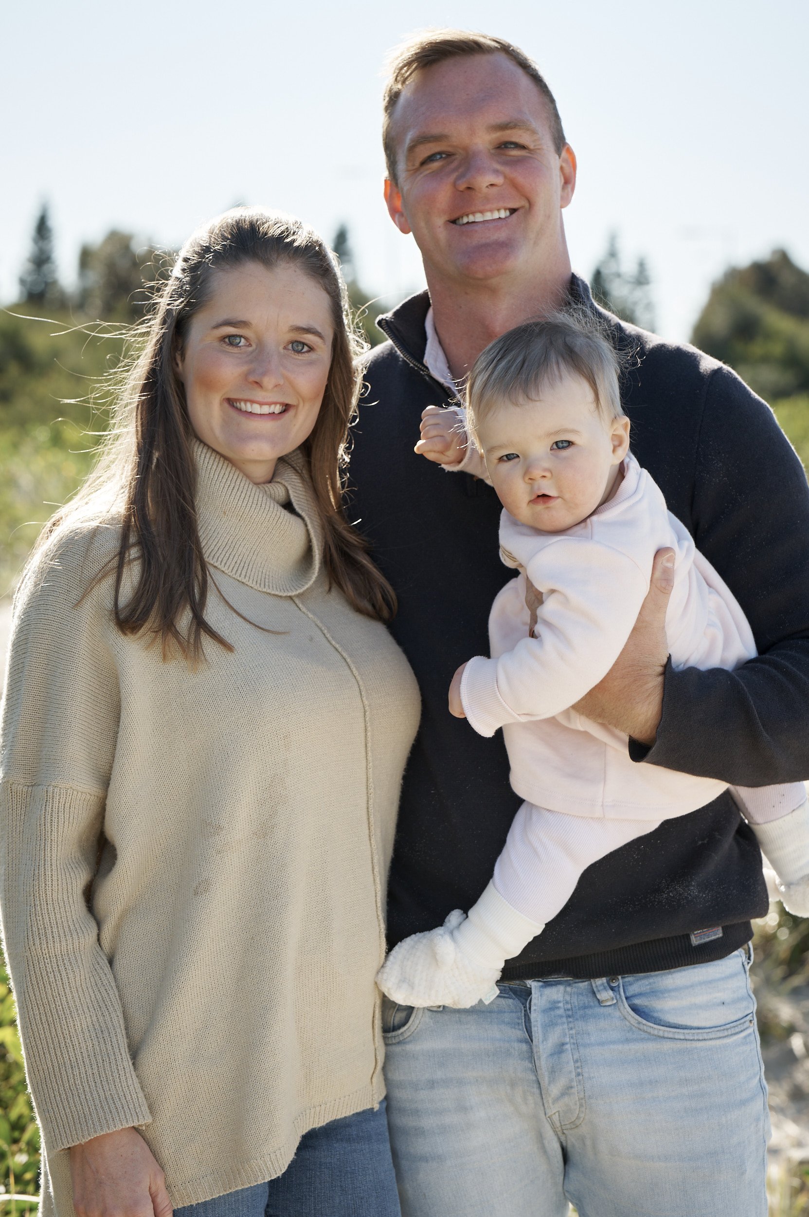 a mother and father stand together smiling at the camera while holding their infant child in their arms