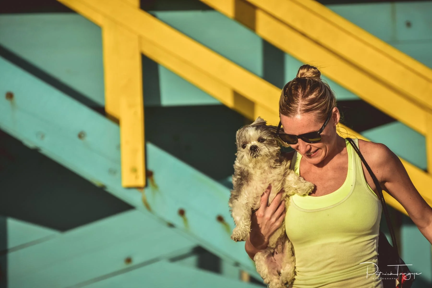 A woman wearing sunglasses and a yellow tank top holding a small fluffy dog, with a colorful wooden staircase in the background.