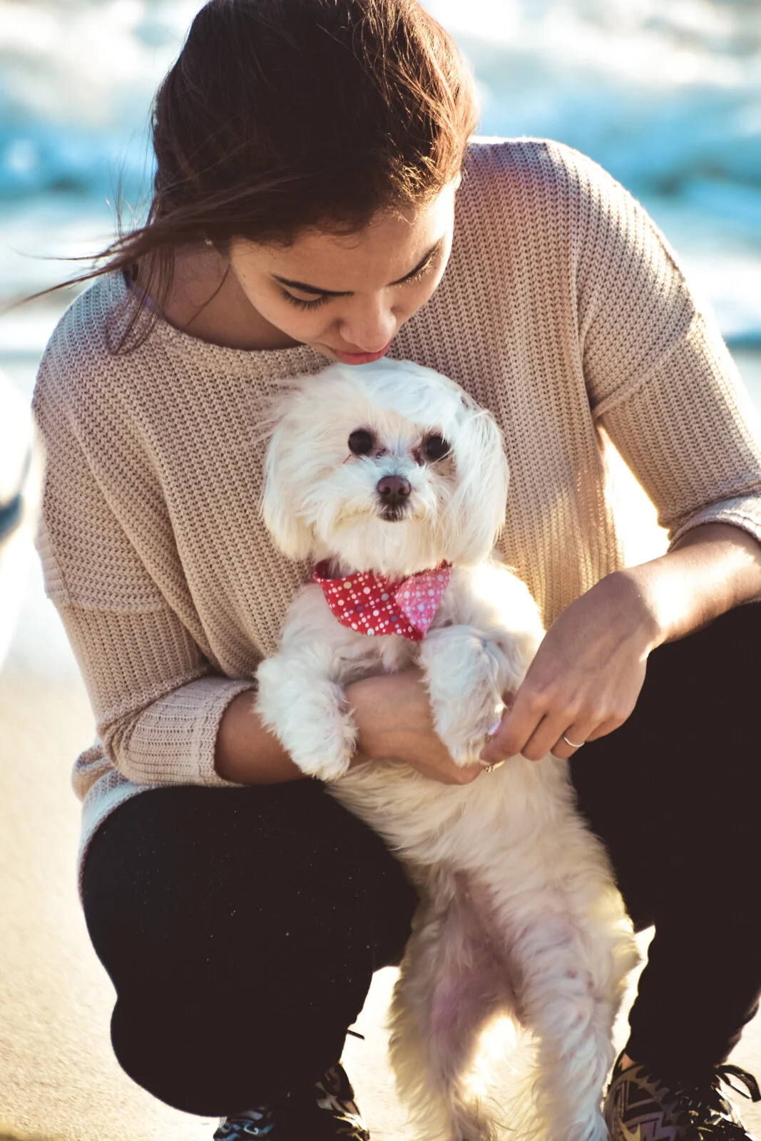 A woman holding a small, white dog with a red polka-dot bandana near a body of water at sunset.